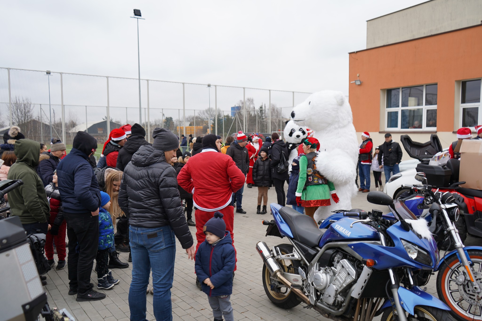 Na zdjęciu grupa ludzi zgromadzonych na zewnątrz na placu szkolnym. W centrum uwagi znajdują się osoby w kostiumach – w tym duży biały niedźwiedź. W tle widoczni są także inni uczestnicy przebrani w czerwonych strojach Mikołajów.