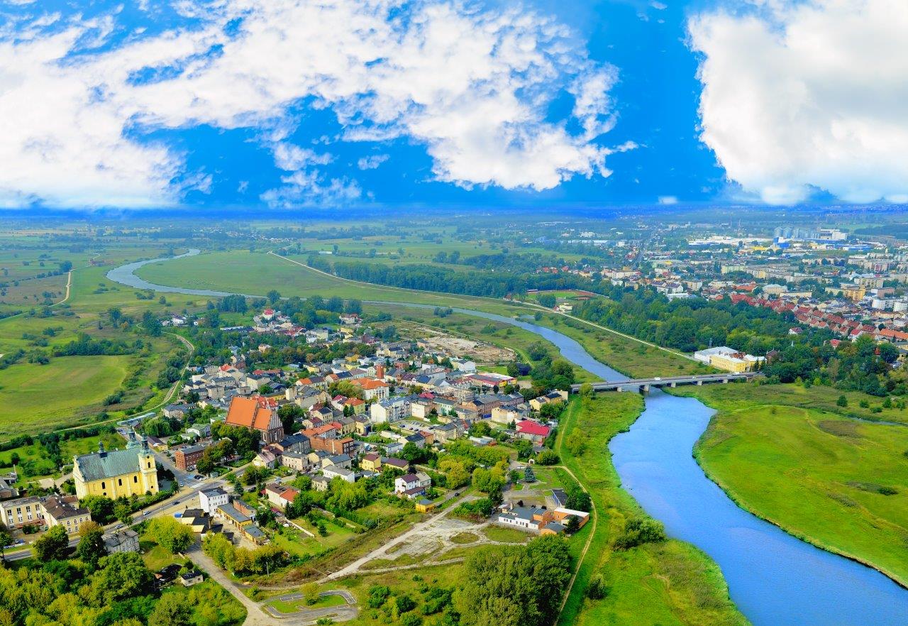 Zdjęcie przedstawia miasto Koło z lotu ptaka, z widocznym zakolem rzeki Warty, mostem, zabudowaniami miejskimi oraz otaczającymi je terenami zielonymi