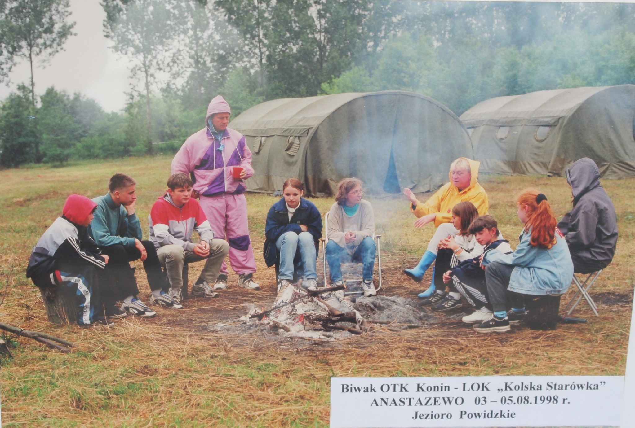 Zdjęcie przedstawia grupę osób siedzących wokół ogniska. W tle widać wojskowe namioty. Uczestnicy obozu są ciepło ubrani. Jeden z dorosłych (osoba w żółtym płaszczu przeciwdeszczowym) prowadzi rozmowę, podczas gdy pozostali słuchają.