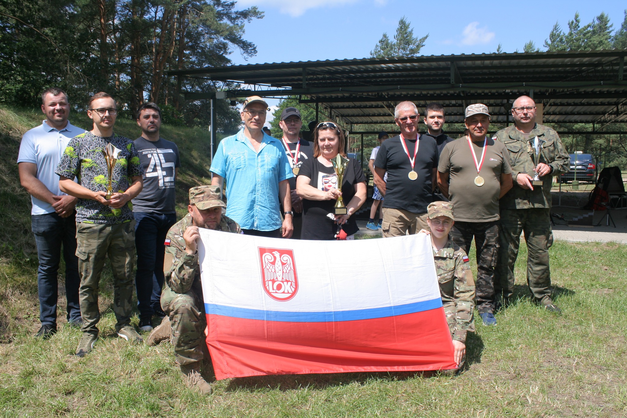 Grupowe zdjęcie uczestników zawodów strzeleckich z pucharami i medalami z flagą biało-czerwoną