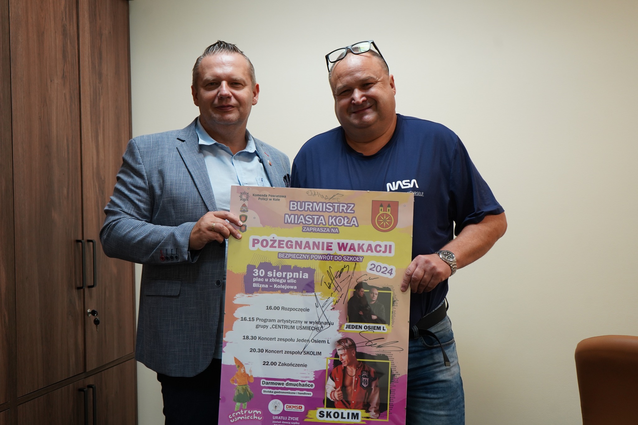 Na zdjęciu znajdują się dwaj mężczyźni, z których jeden trzyma plakat promujący wydarzenie 