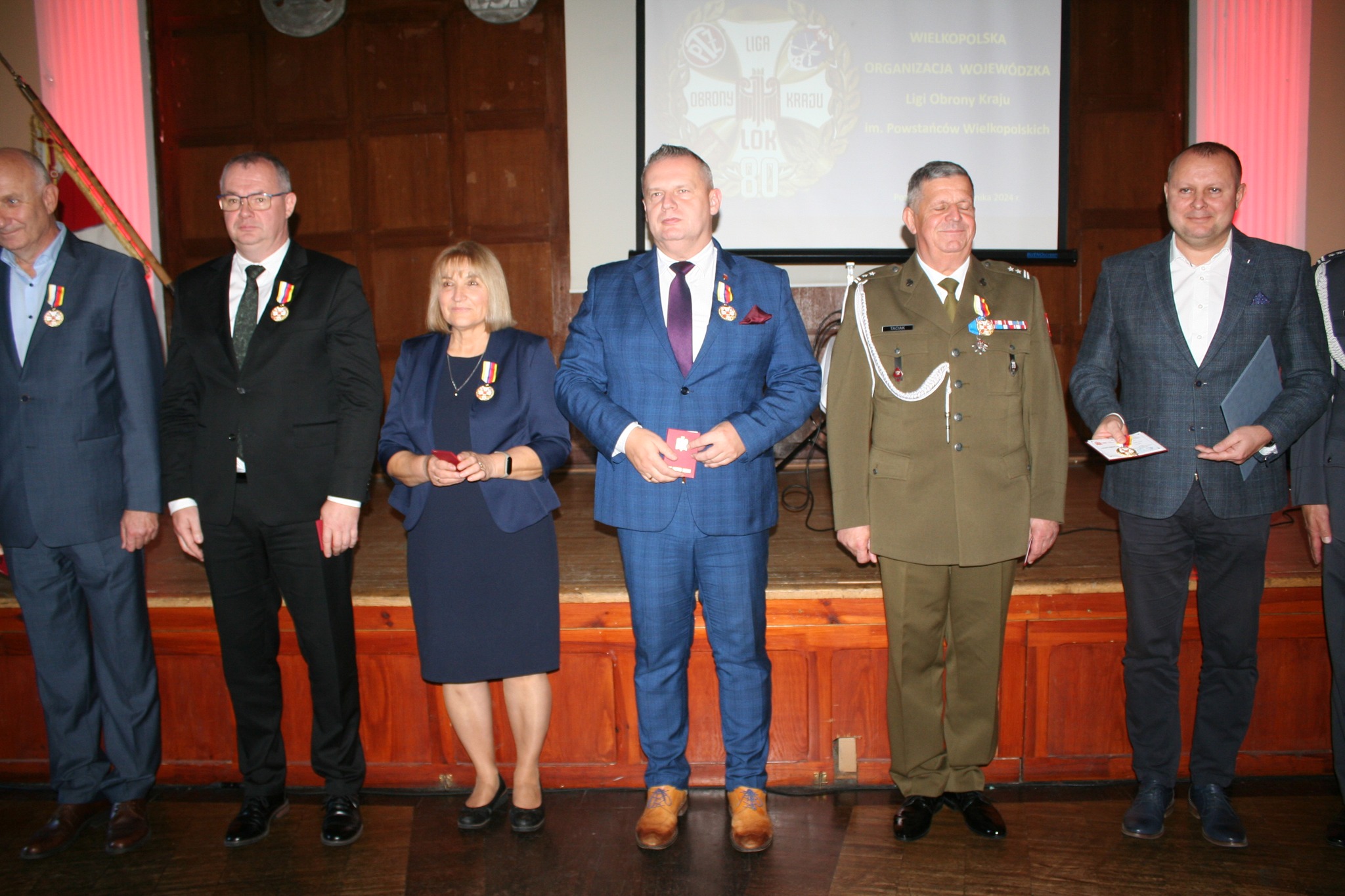 Na zdjęciu widać sześć stojących osób. Każda z nich ma przypięty medal na piersi. Pośrodku znajduje się mężczyzna w niebieskim garniturze, obok niego kobieta w granatowej garsonce oraz mężczyzna w mundurze wojskowym. 