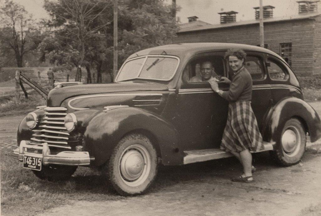 Na fotografii widzimy stary samochód z lat 50. XX wieku. Przy samochodzie stoi kobieta w spódnicy w kratę, oparta o drzwi pasażera, a wewnątrz samochodu siedzi mężczyzna, patrzący przez otwarte okno. Zdjęcie wykonane w wiejskiej scenerii.