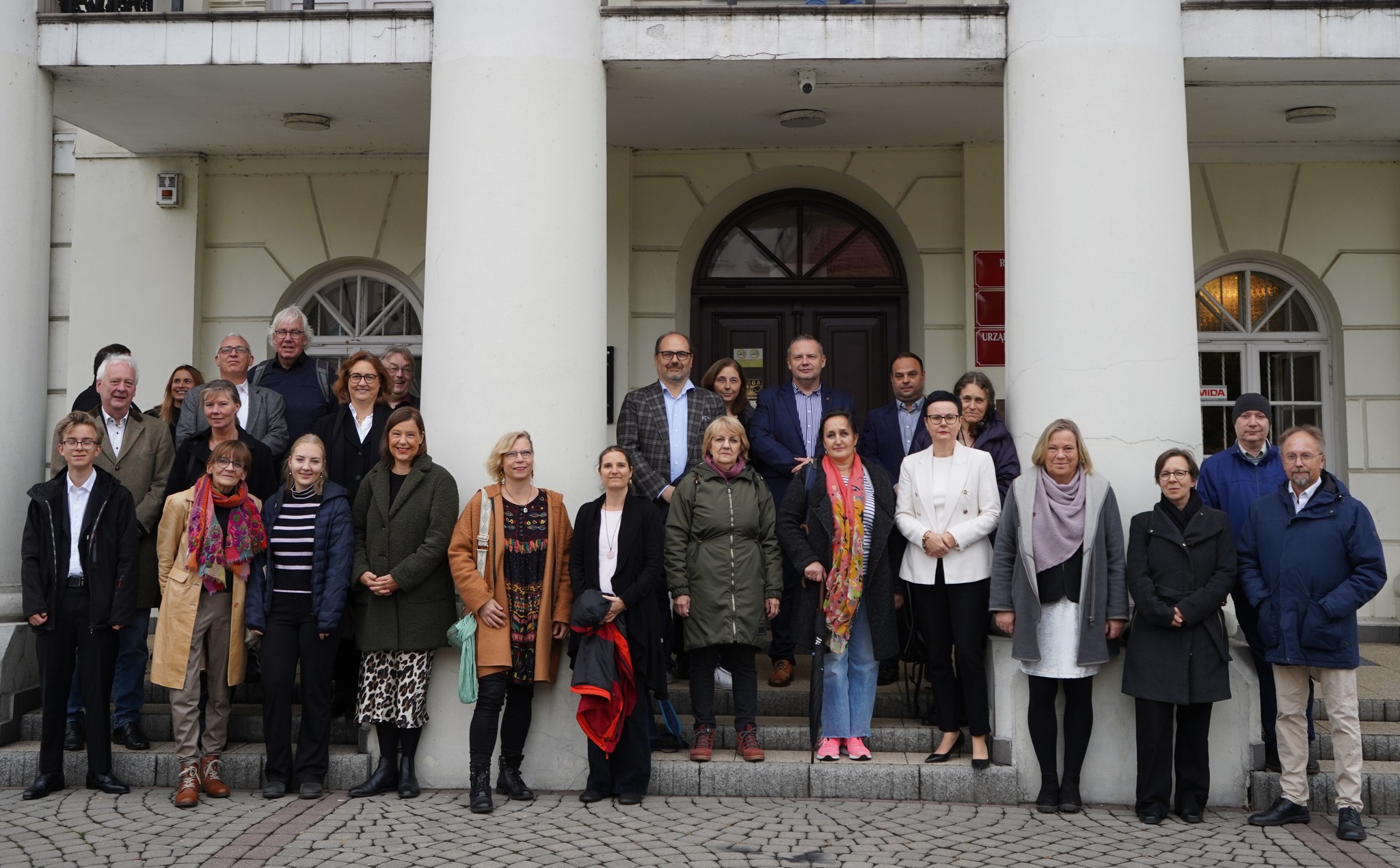 Na zdjęciu grupa osób stojących na schodach przed wejściem do Ratusza. Grupa składa się z mężczyzn i kobiet, wszyscy są ubrani na jesienno-zimowo. W tle widoczne są drzwi oraz fragment budynku z łukowymi oknami.