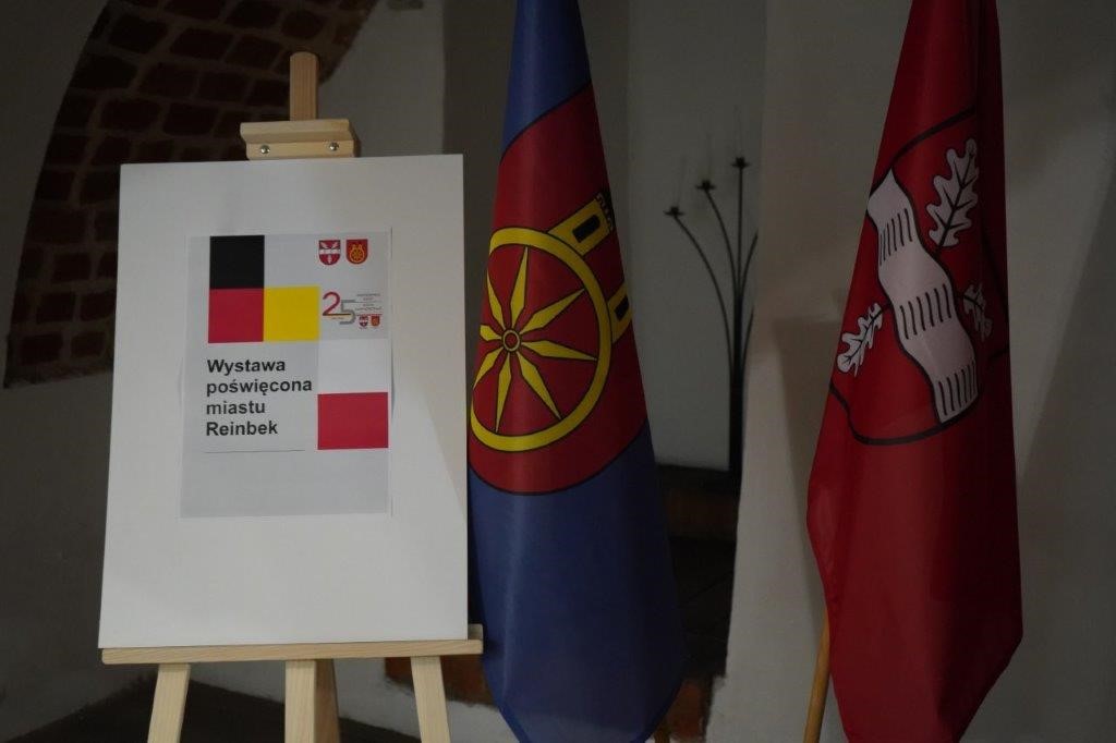 Flagi miasta partnerskiego Reinbek i Miasta Koła, obok sztaluga z tablicą informującą o wystawie