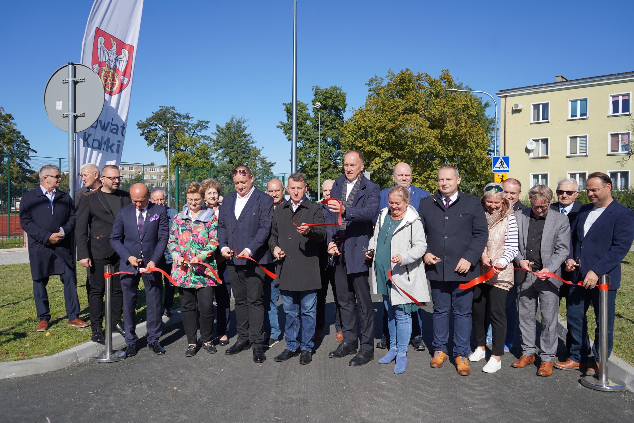 Na zdjęciu widać grupę osób, która bierze udział w ceremonii przecięcia wstęgi, co oznacza otwarcie przebudowanej ulicy Broniewskiego w Kole. W tle widać bloki mieszkalne oraz flagę z napisem Powiat Kolski