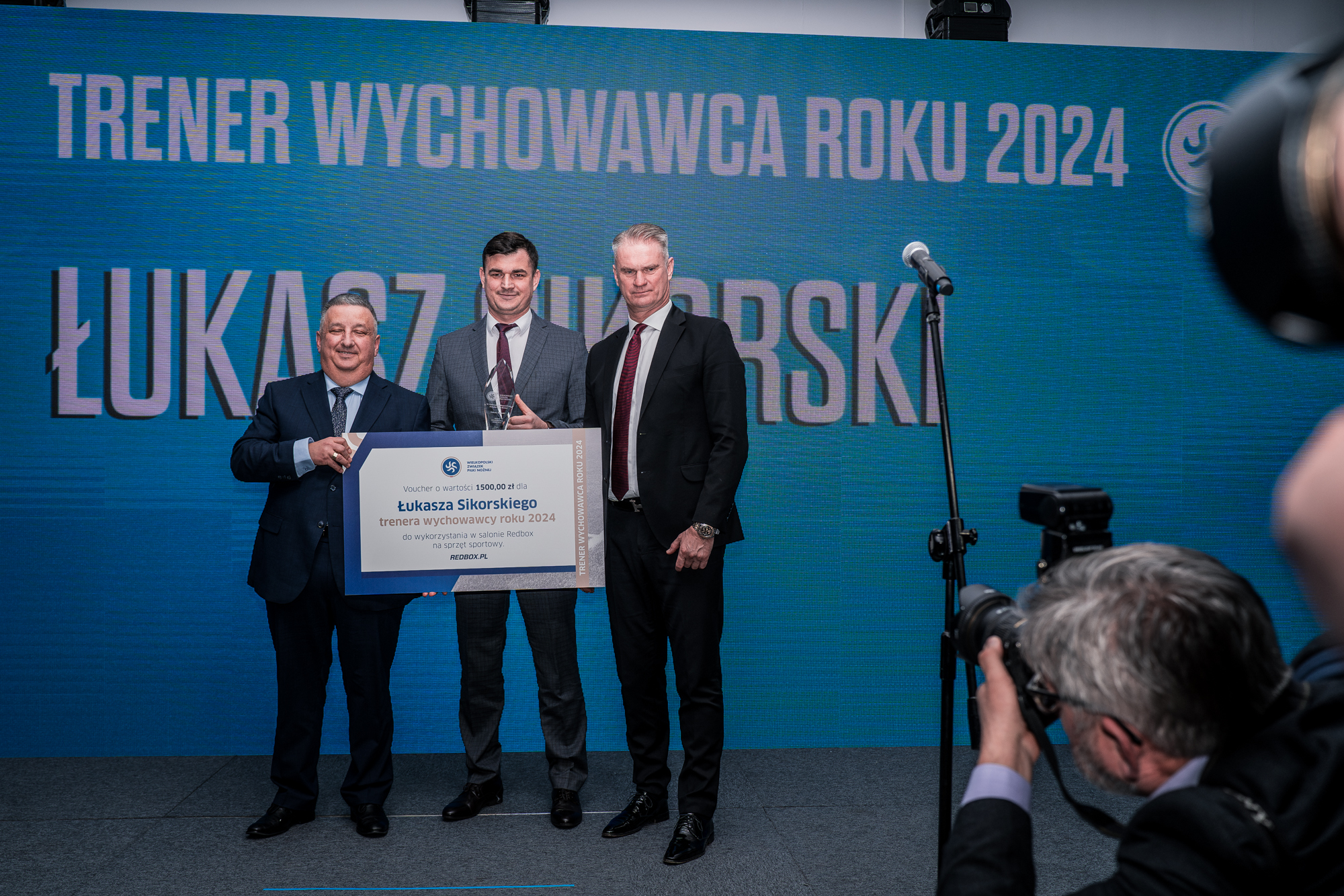 Na zdjęciu widać trzech eleganckich mężczyzn stojących na ceremonii wręczenia nagród. W tle napis Trener Wychowawca Roku 2024 oraz nazwisko laureata, Łukasza Sikorskiego. Jeden z mężczyzn trzyma pamiątkową tablicę z voucherem i statuetkę.