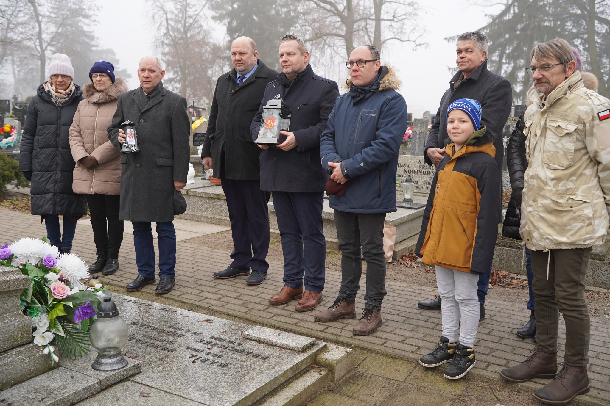 Na zdjęciu grupa osób stoi przy grobie na cmentarzu, trzymając znicze.