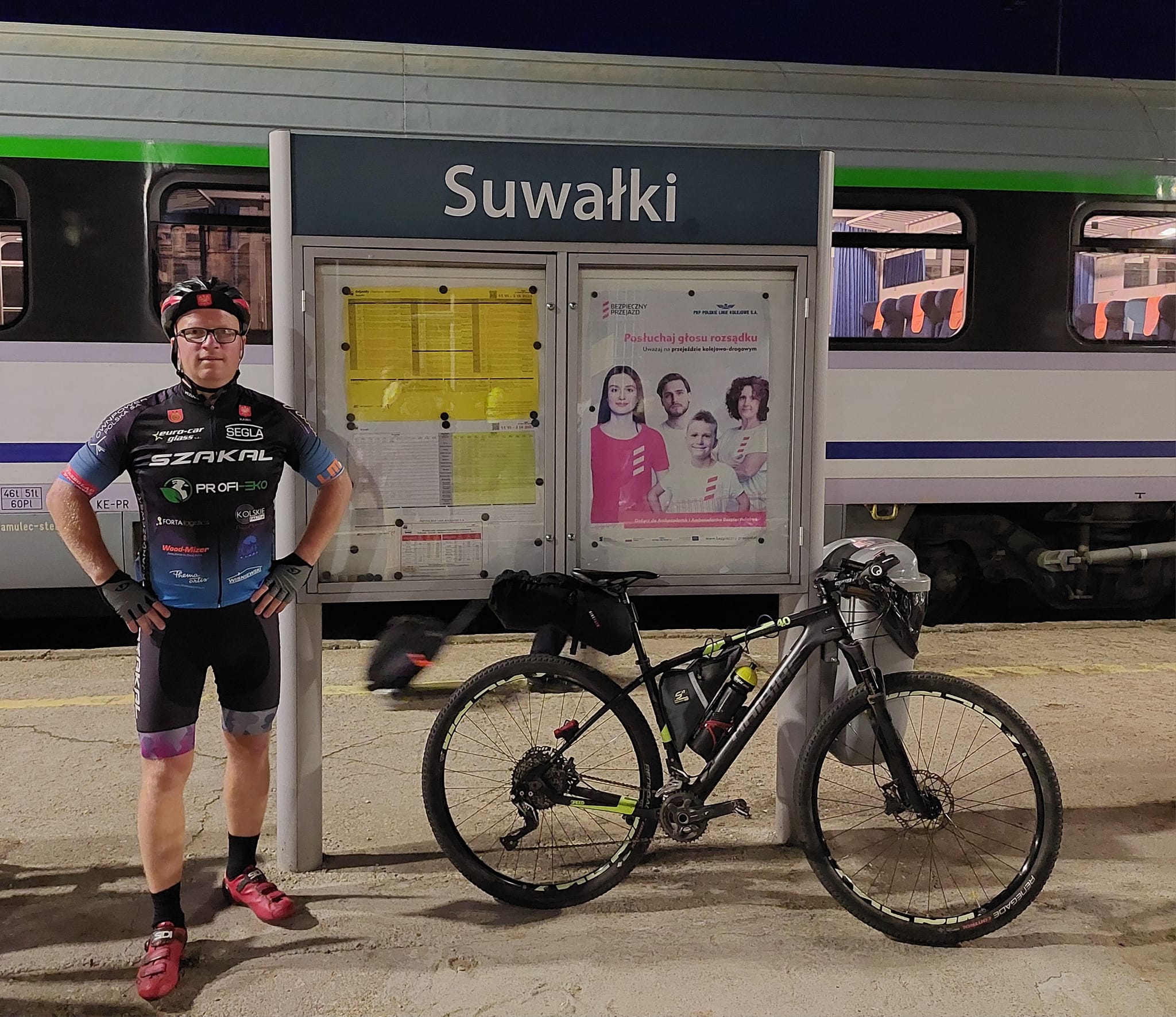 Kamil Jaroszewski stoi z rowerem na peronie na stacji przy znaku z napisem Suwałki