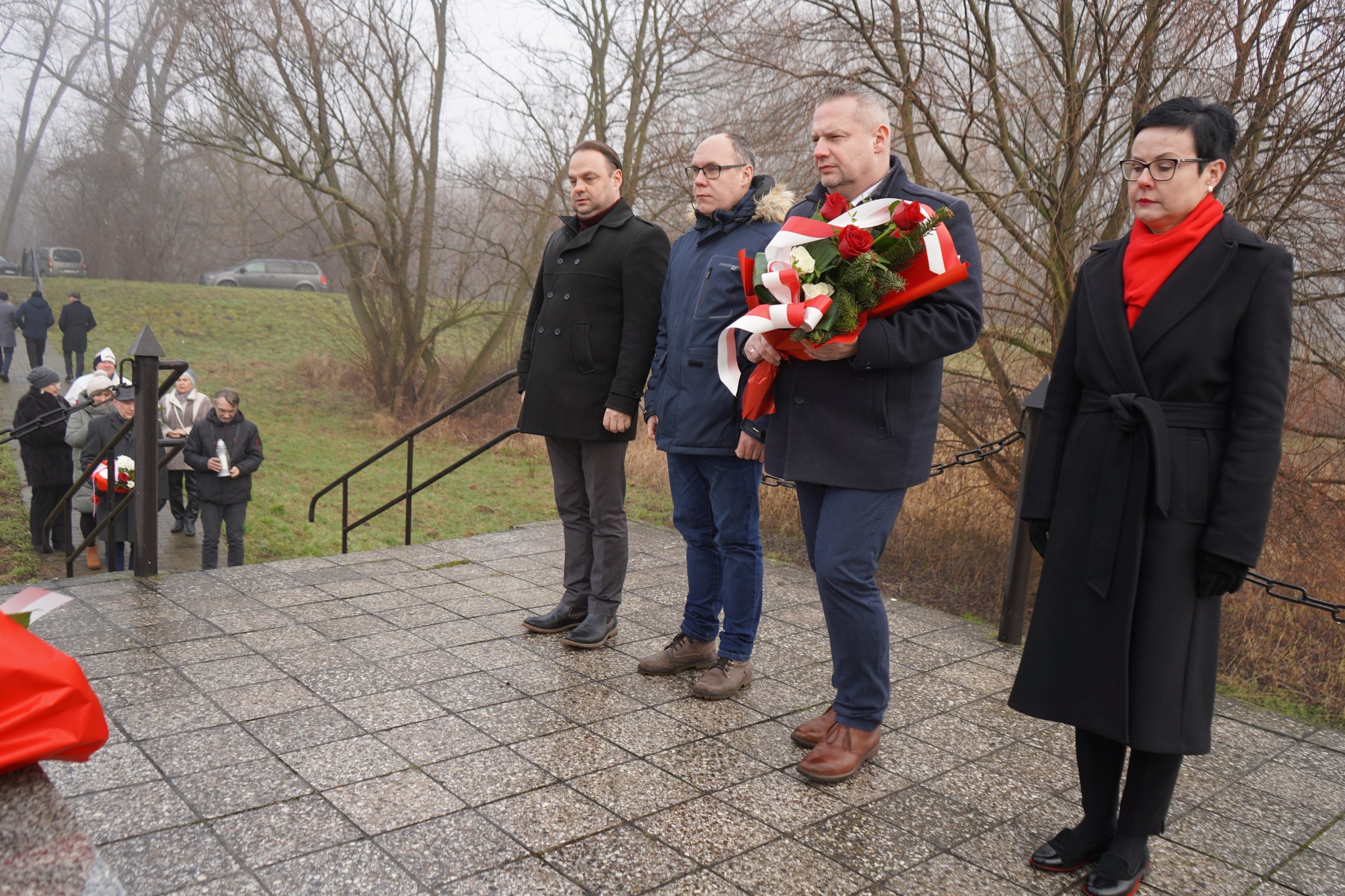 Na zdjęciu widać cztery osoby przed pomnikiem podczas obchodów Rocznicy Powstania Wielkopolskiego w Kole. Jedna z osób trzyma wieniec z biało-czerwonymi wstęgami. W tle występują inni uczestnicy wydarzenia oraz zimowy, bezlistny krajobraz. 