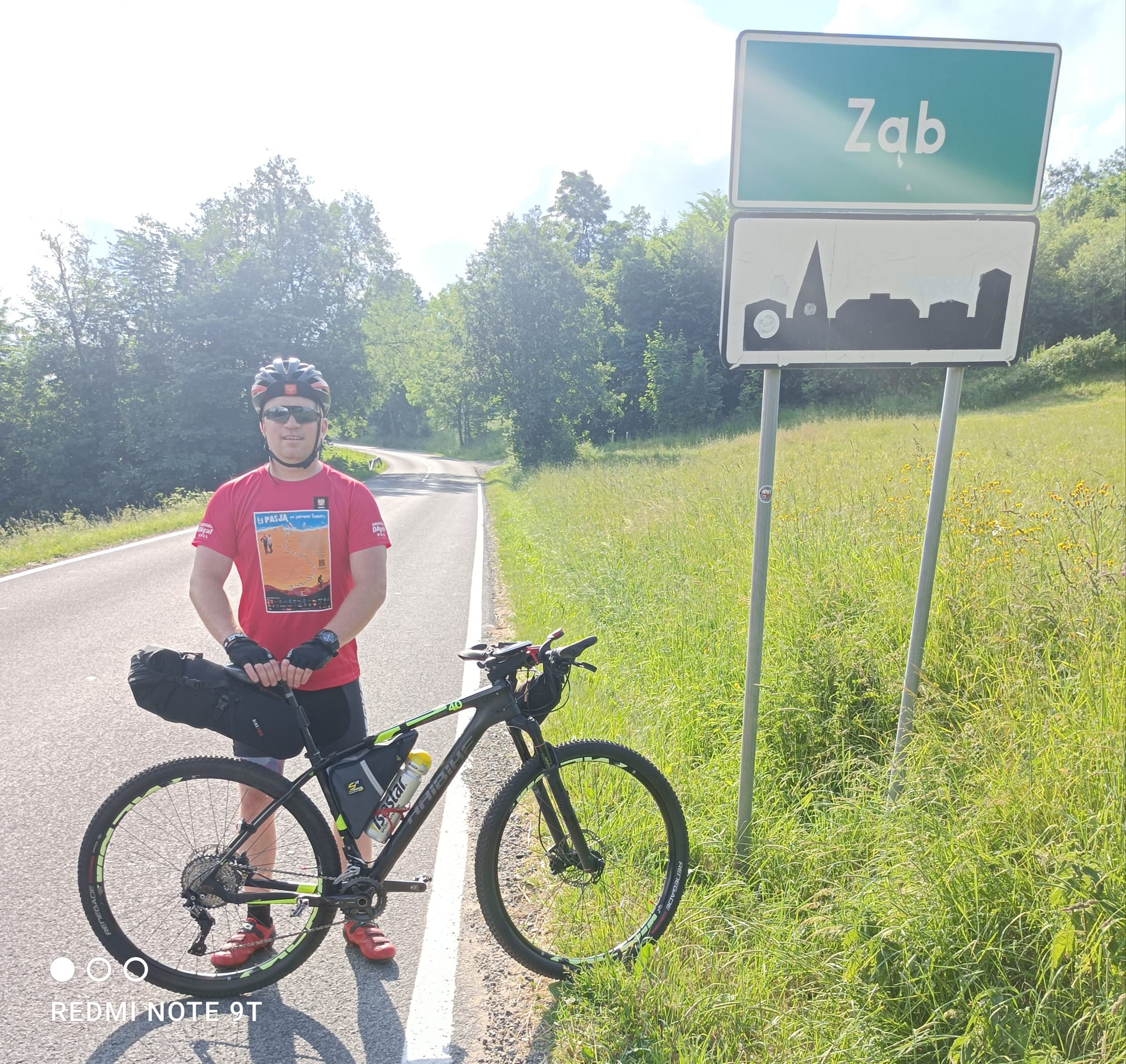 Kamil Jaroszewski stoi z rowerem na tle znaku miejscowości Ząb.