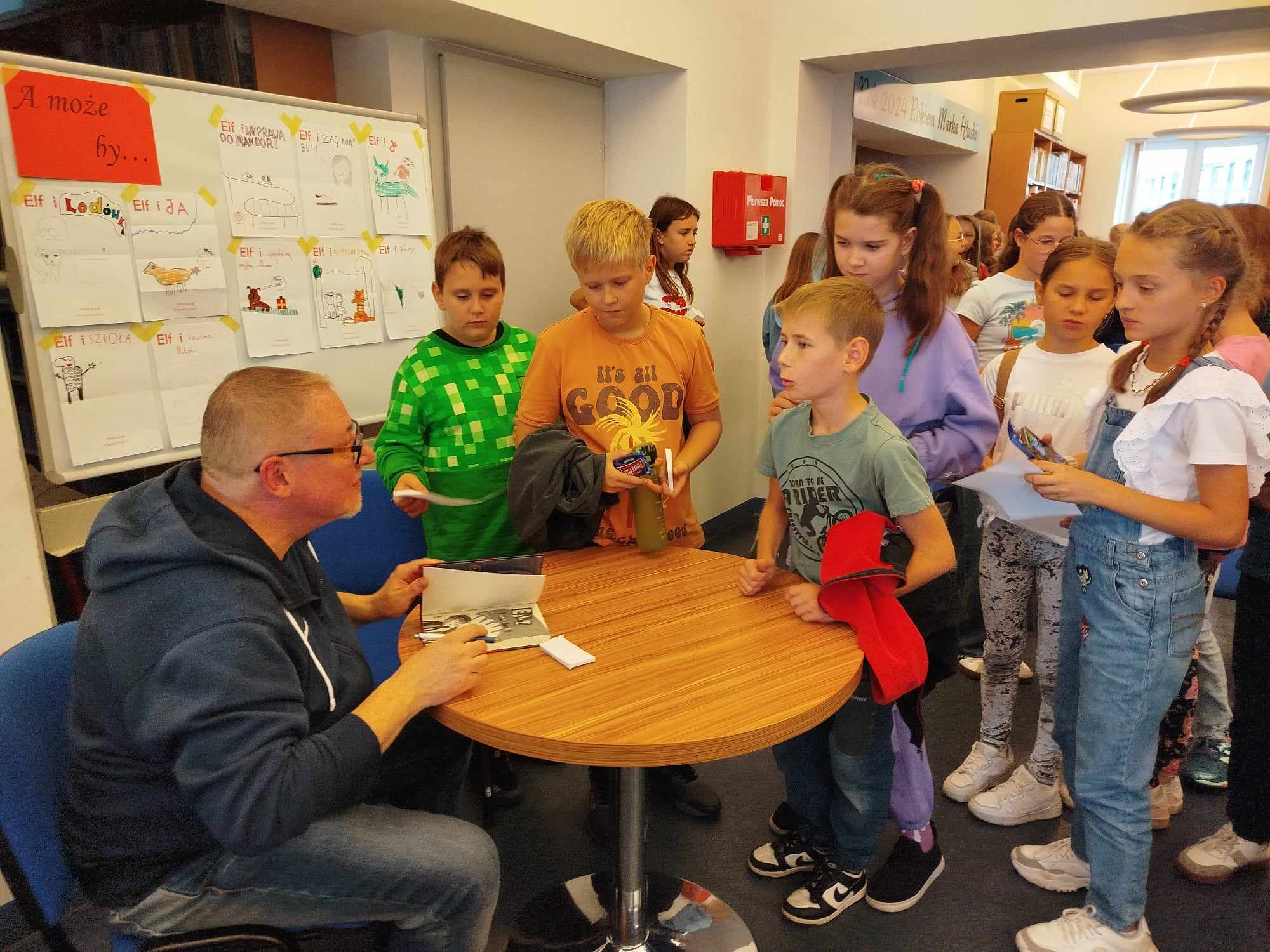 Zdjęcie przedstawia grupę dzieci stojących w kolejce do pisarza siedzącego przy stole. Mężczyzna podpisuje książkę, W tle na tablicy znajdują się rysunki, z napisami zaczynającymi się od ELF. Dzieci mają na sobie codzienne ubrania, są uśmiechnięci.