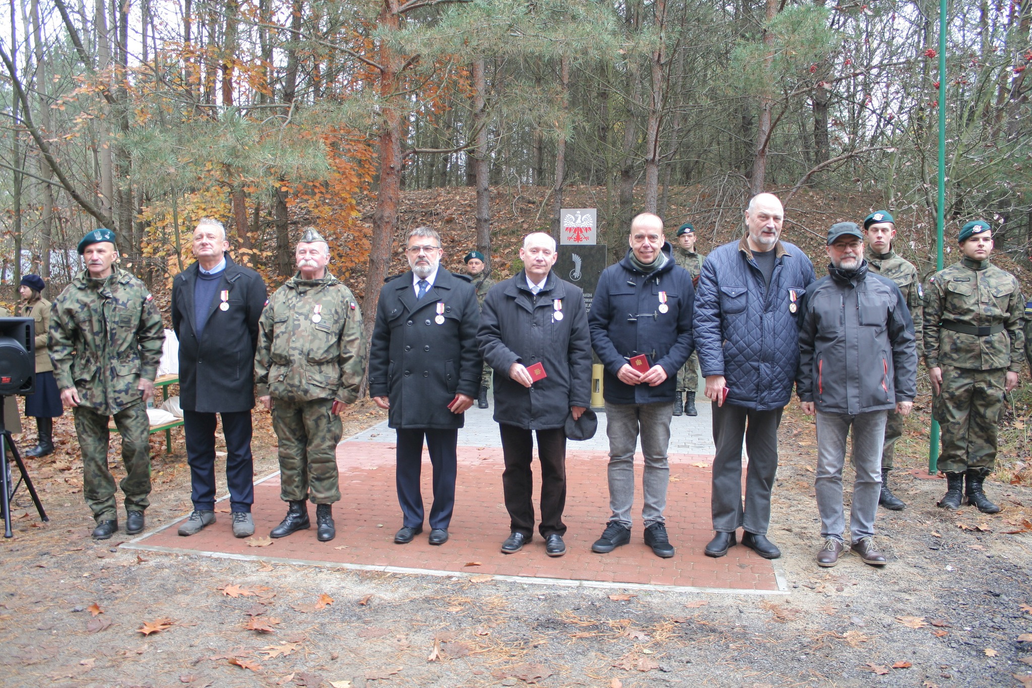 Zdjęcie przedstawia grupę mężczyzn stojących w szeregu, z odznaczeniami przypiętymi do ubrań. Za nimi znajduje się las w jesiennych kolorach, a w tle widoczny jest pomnik.