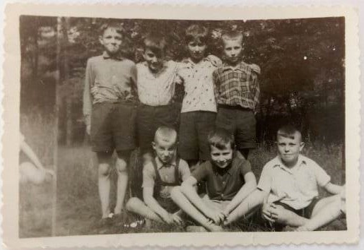 Zdjęcie przedstawia grupę siedmiu chłopców w 1961 r. Są ubrani w krótkie spodenki i koszule, stoją na tle drzew. Trzech stoi z tyłu, a trzech siedzi na ziemi z przodu. Zdjęcie ma charakterystyczne ząbkowane krawędzie, typowe dla starych fotografii.