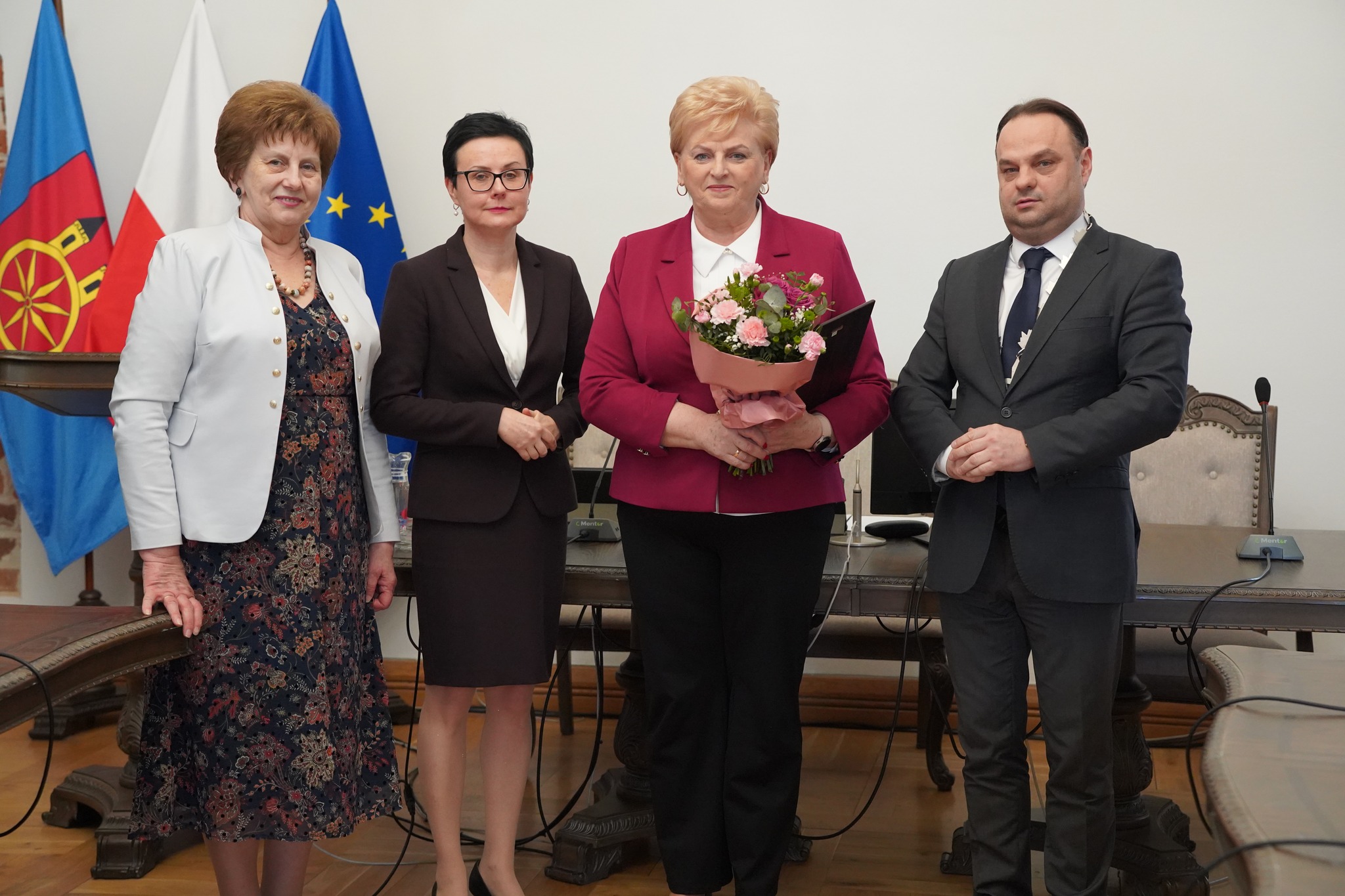 Na zdjęciu widać cztery osoby w sali sesyjnej Ratusza w Kole. Trzy kobiety z lewej i mężczyznę z prawej strony. Kobieta w bordowej marynarce trzyma bukiet kwiatów i dyplom. W tle znajduje się flaga Polski, Unii Europejskiej i samorządu. miasta Koła.