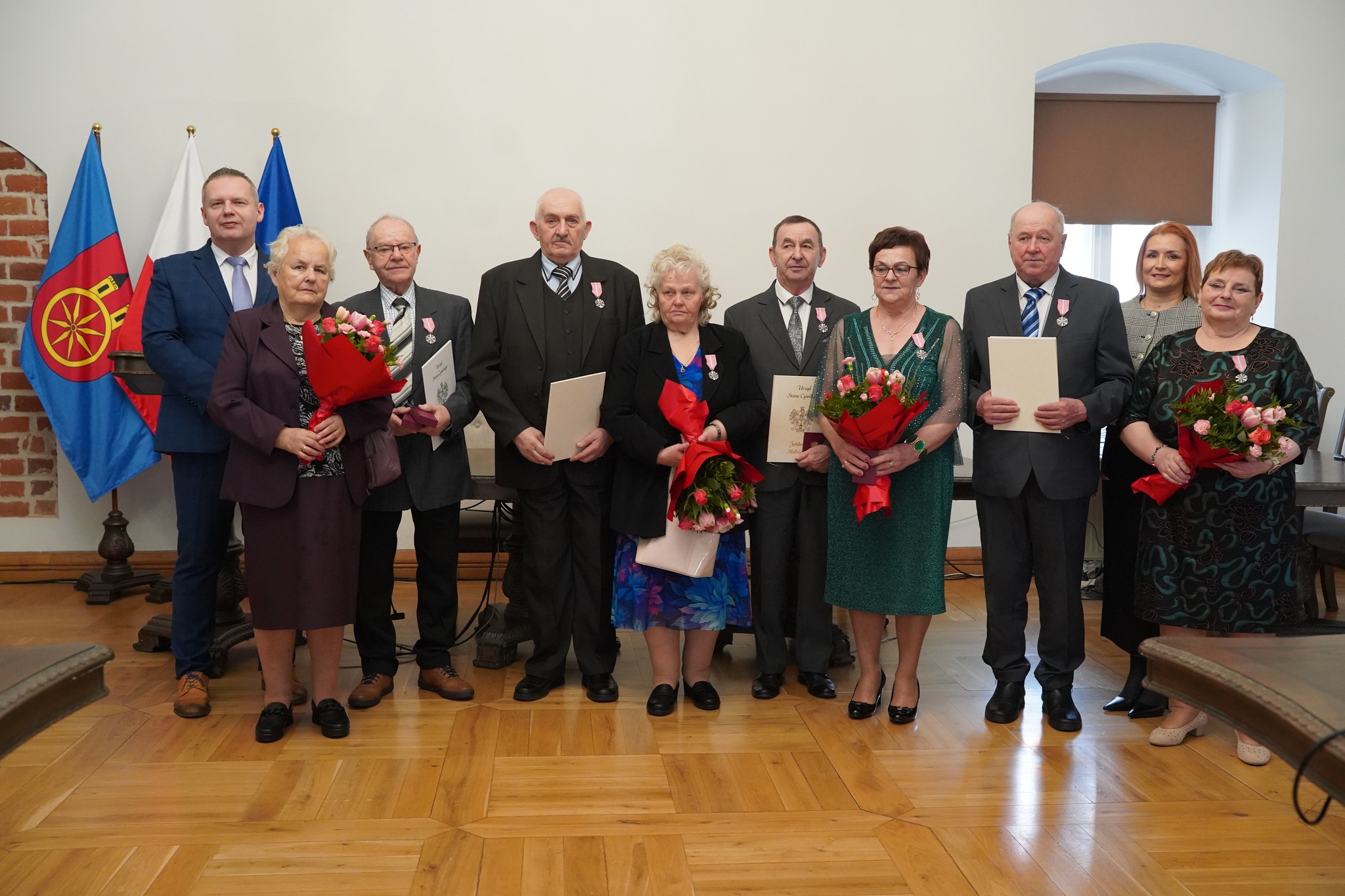 Na zdjęciu widać grupę elegancko ubranych osób, które trzymają odznaczenia i kwiaty z okazji jubileuszu pożycia małżeńskiego. W tle widoczne są flagi oraz oficjalne dokumenty, co podkreśla uroczysty charakter wydarzenia.