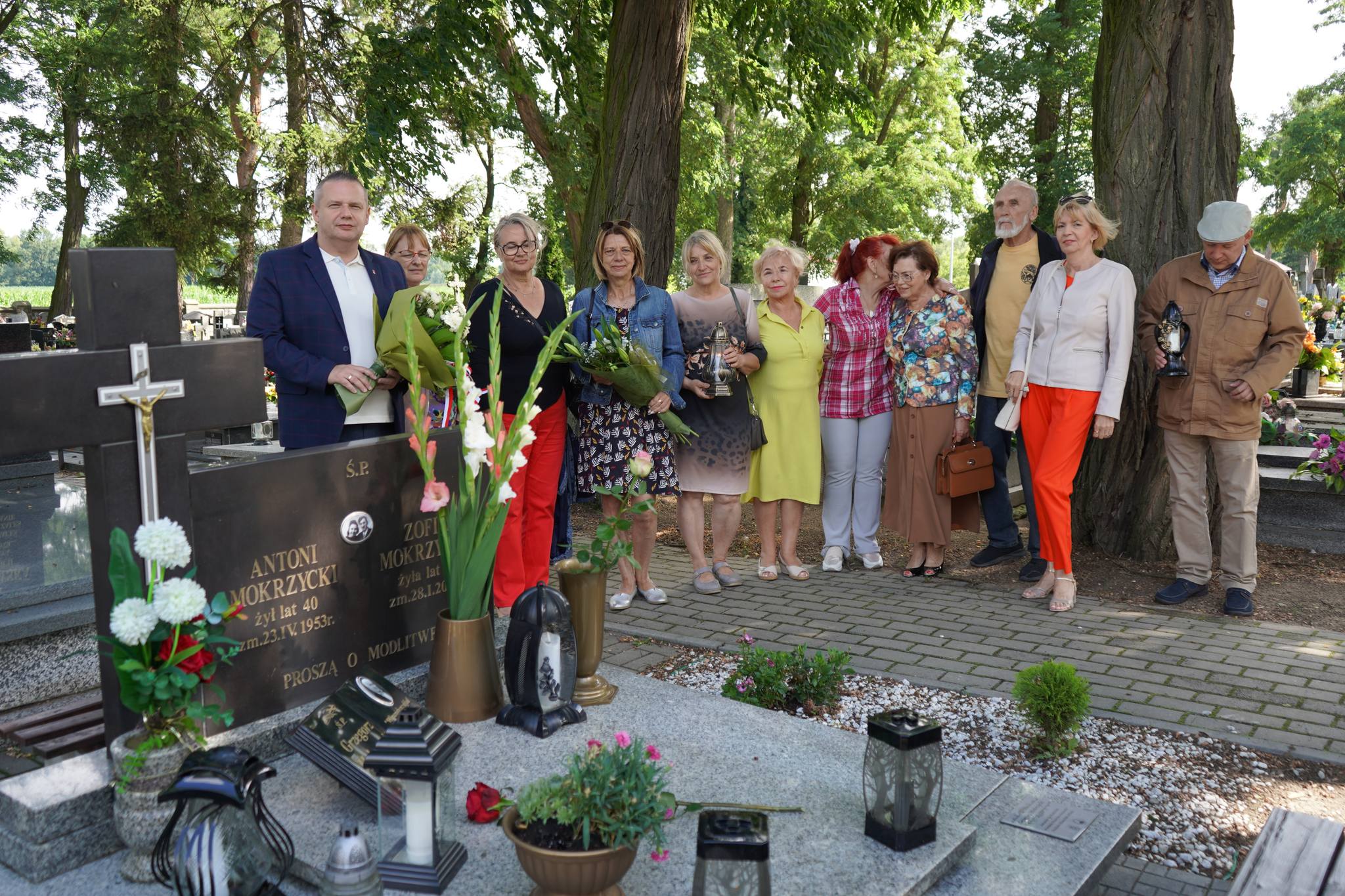 Zdjęcie przedstawia grupę ludzi zgromadzonych na cmentarzu, w tle widać zielone drzewa, a grupa osób trzyma kwiaty i znicze. Atmosfera jest spokojna i pełna zadumy, jest okazją upamiętnienia zmarłego  Grzegorza Mokrzyckiego