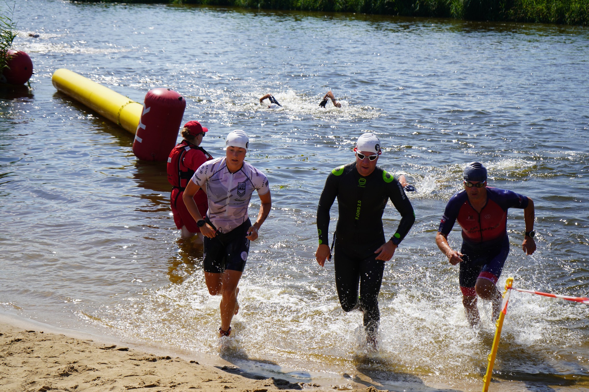 Na zdjęciu widać uczestników triathlonu wychodzących z wody na brzeg. Trzech zawodników w strojach pływackich i czepkach śpieszy się w kierunku linii wyznaczonej przez taśmę. W tle widać inne osoby w wodzie, a także wolontariusza w czerwonej kamizelc