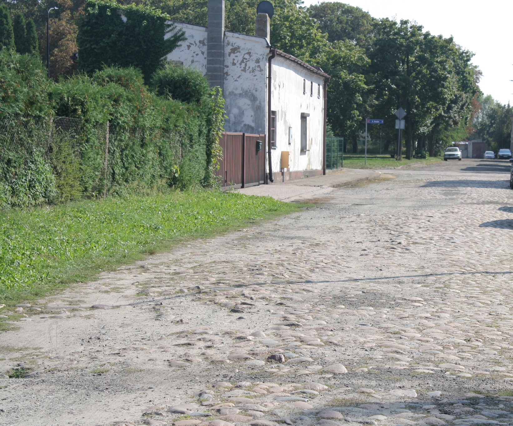 Zdjęcie przedstawia brukowaną drogę, z lewej strony widoczny jest pas zieleni, za nim stoi stary budynek z bramą, natomiast w tle widać drzewa i jadący samochód