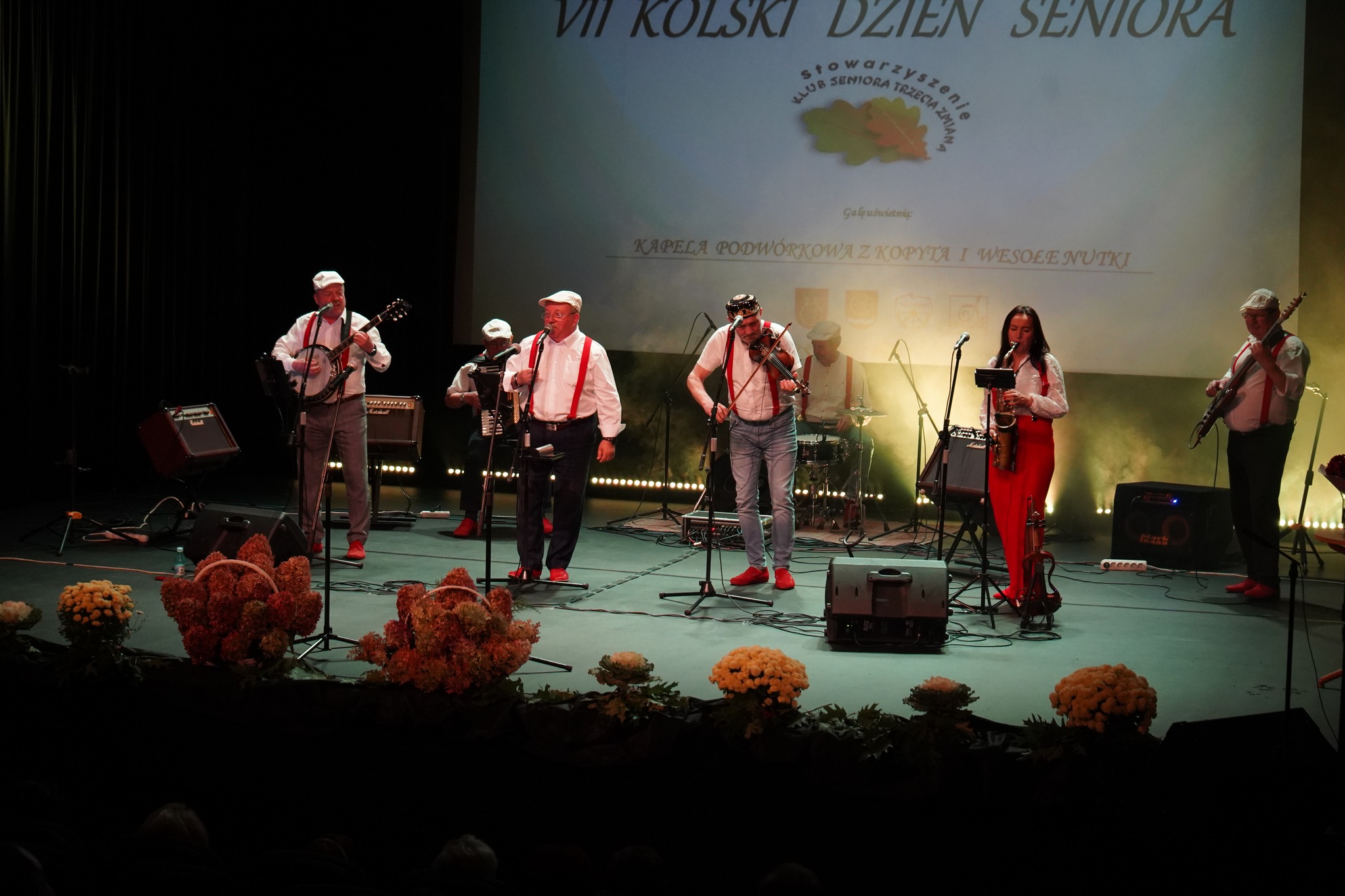 Zdjęcie przedstawia kapelę podwórkową grającą na scenie z okazji Dnia Seniora. Muzycy są ubrani w czerwone szelki oraz czerwone buty. Grają na saksofonie, skrzypcach, i banjo. Tło stanowi ekran z napisem VII Kolski Dzień Seniora