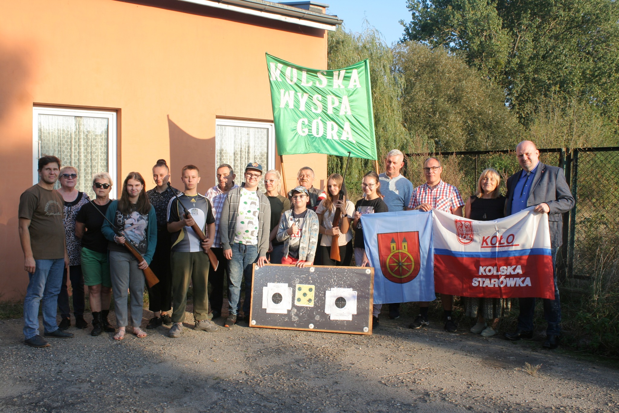 Zdjęcie przedstawia grupę młodych ludzi oraz dorosłych stojących na zewnątrz przed budynkiem. Na pierwszym planie, kilka osób trzyma flagi z napisami KOŁO, KOLSKA STARÓWKA i KOLSKA WYSPA GÓRĄ. Kilkoro dzieci trzyma w dłoniach karabinki pneumatyczne.