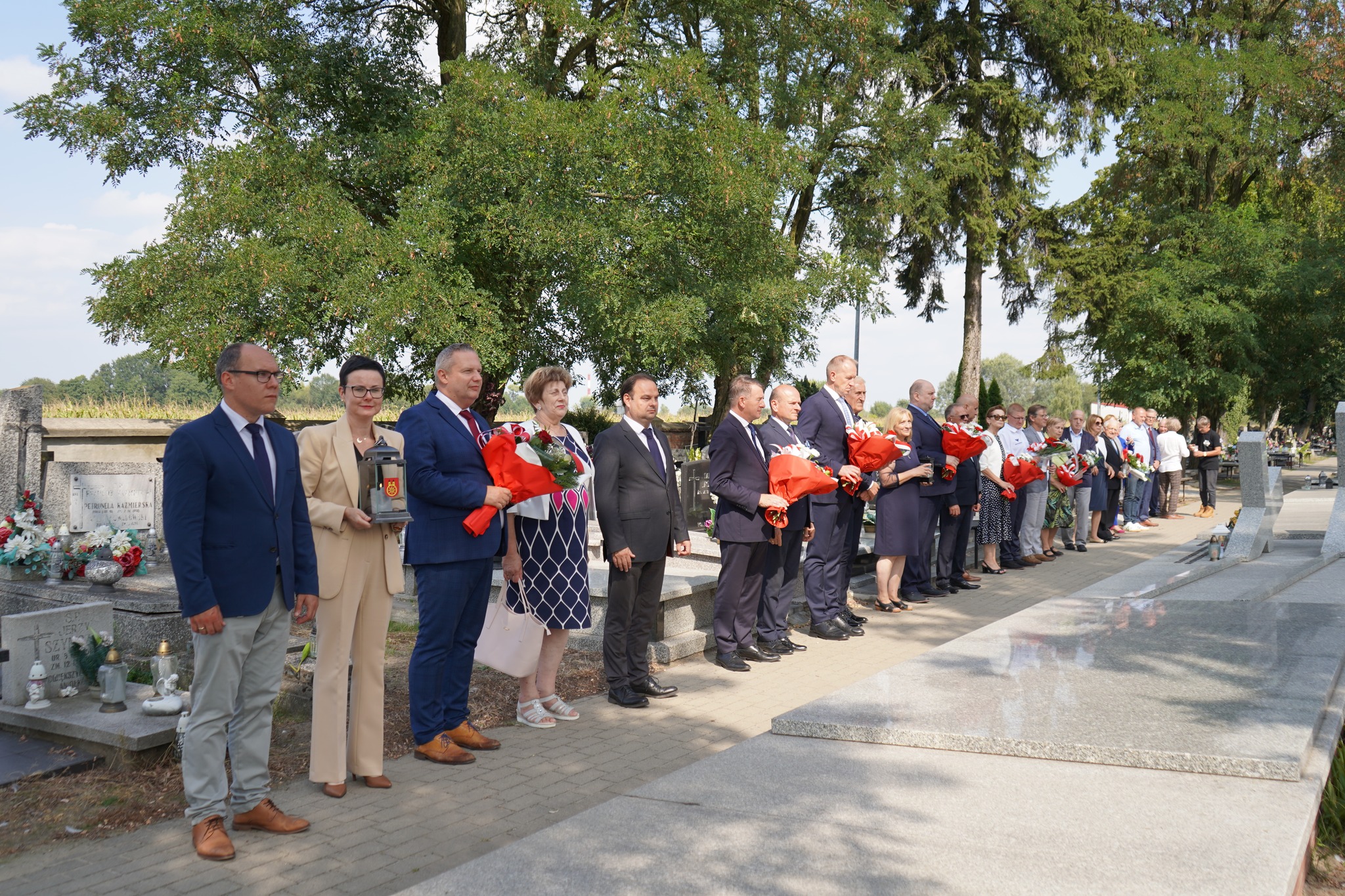 Zdjęcie przedstawia grupę dorosłych osób zebranych przy zbiorowej mogile ofiar na cmentarzu parafialnym w Kole. Grupa liczy kilkanaście osób, kilka trzyma w dłoniach wiązanki kwiatów. W tle widać zielone drzewa.