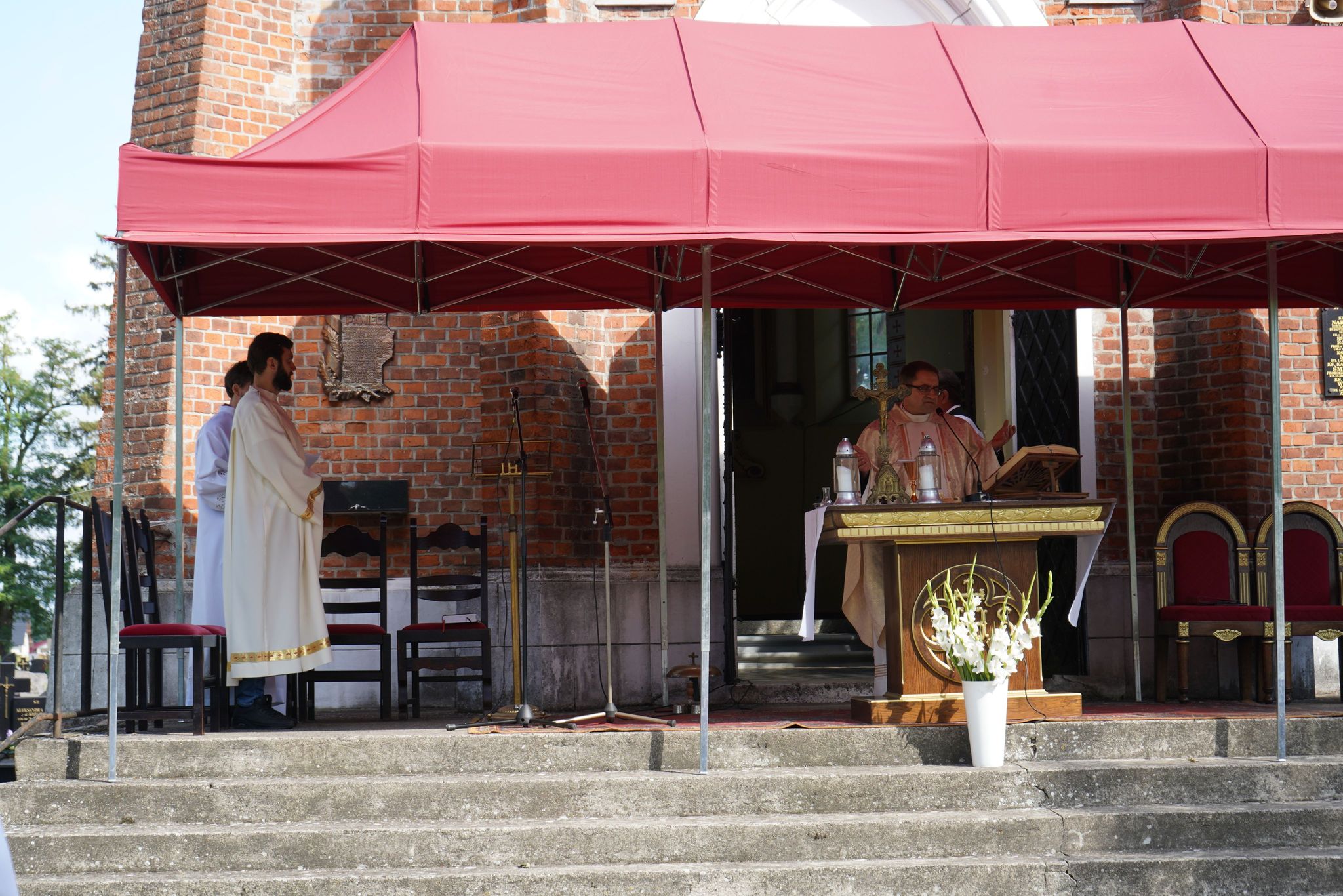 Na zdjęciu widzimy scenę z ceremonii religijnej odbywającej się na zewnątrz, przed kaplicą z czerwonej cegły na cmentarzu parafialnym w Kole. Na podwyższeniu znajduje się dwóch duchownych ubranych w liturgiczne szaty.