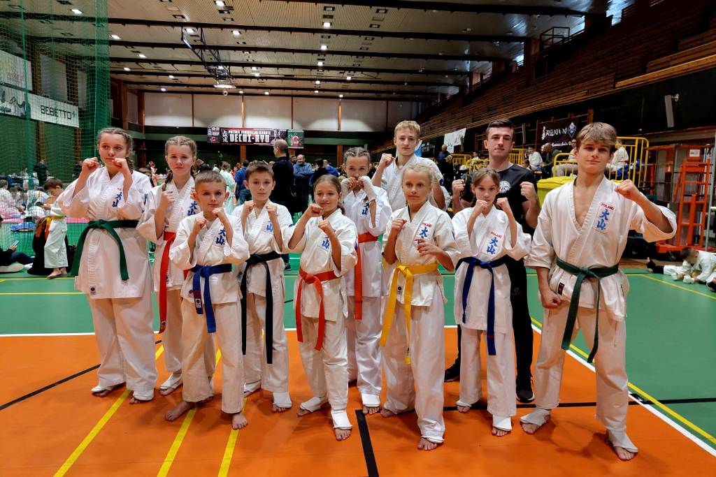 Na zdjęciu widać grupę młodych zawodniczek i zawodników Oyama Karate stojących w strojach kimono z podniesionymi dłońmi z zaciśniętymi w pięści w hali sportowej. W tle, z prawej strony, widać trybuny, z lewej natomiast zielone siatki zabezpieczające