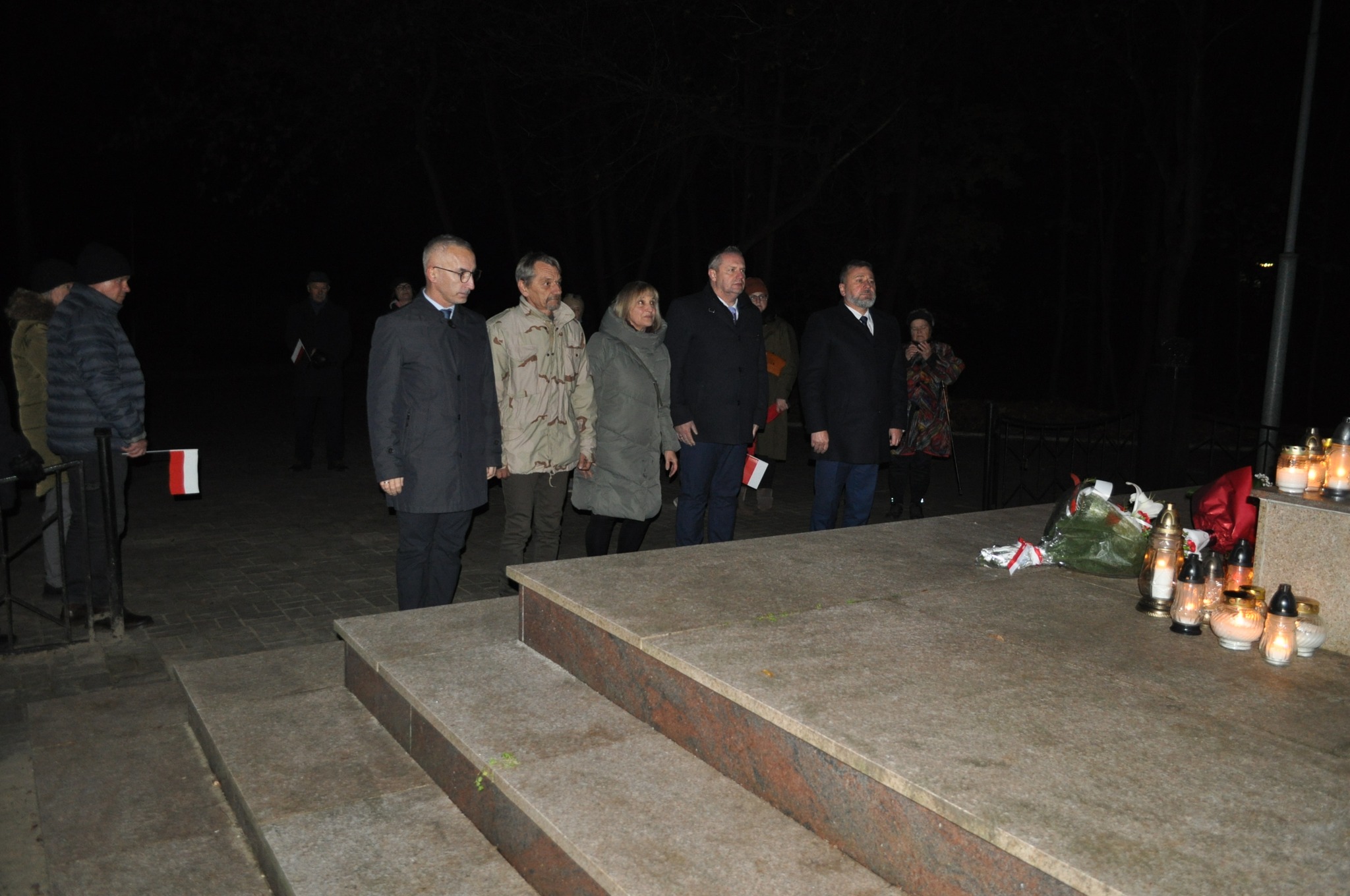 Na zdjęciu widać grupę osób stojących w ciszy przed pomnikiem. W tle są znicze oraz kwiaty, co sugeruje, że oddają hołd lub składają wyrazy pamięci.