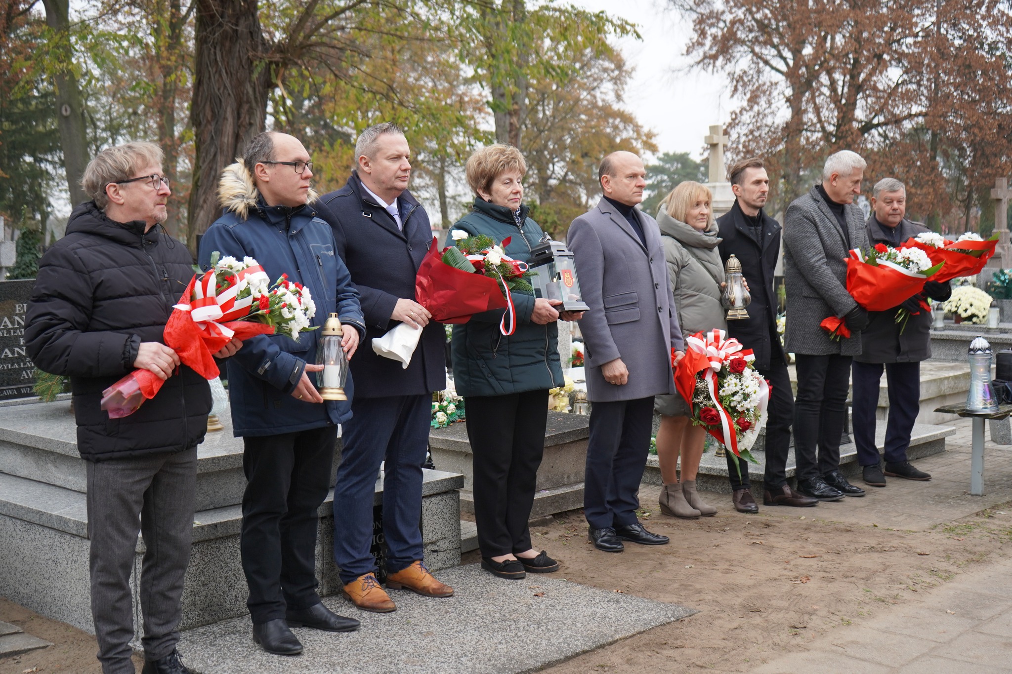 Na zdjęciu widzimy grupę dziewięciu osób stojących przy grobie na cmentarzu, biorących udział w ceremonii upamiętniającej Czesława i Krystyny Freudenreichów. Każda z osób trzyma kwiaty lub znicze. W tle znajdują się groby oraz drzewa.