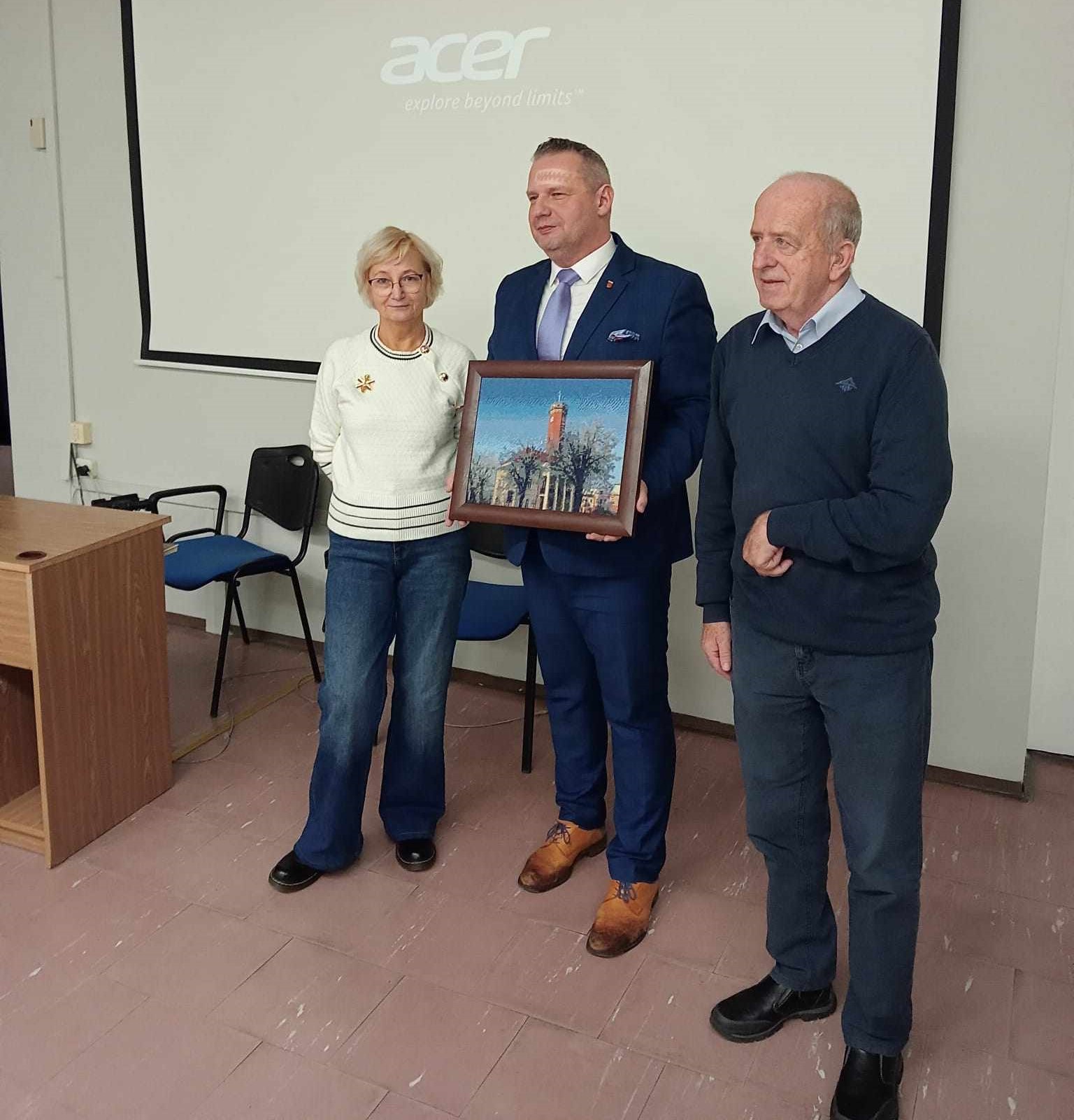 Zdjęcie przedstawia trzy osoby w sali konferencyjnej liceum ogólnokształcącego w Kole. Środkowa osoba trzyma oprawiony obraz z widokiem na budynek Ratusza w Kole. W tle widoczny jest ekran projekcyjny.