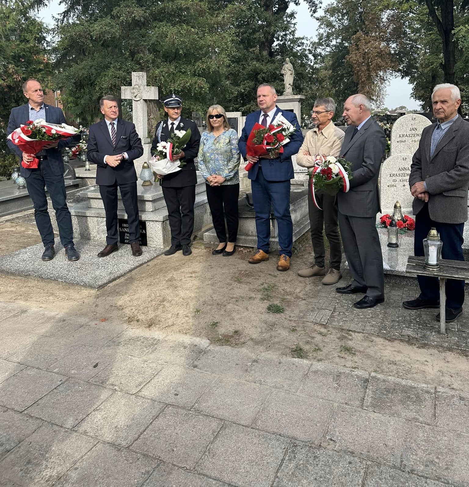 Zdjęcie przedstawia grupę ludzi zgromadzonych na cmentarzu. W tle widać zielone drzewa, a grupa osób trzyma kwiaty i znicze.