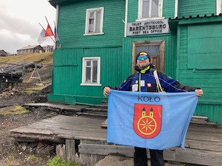 Zdjęcie przedstawia osobę trzymającą flagę z herbem miasta Koło, stojącą przed zielonym budynkiem oznaczonym napisem „Barentsburg Port Sea Office”.