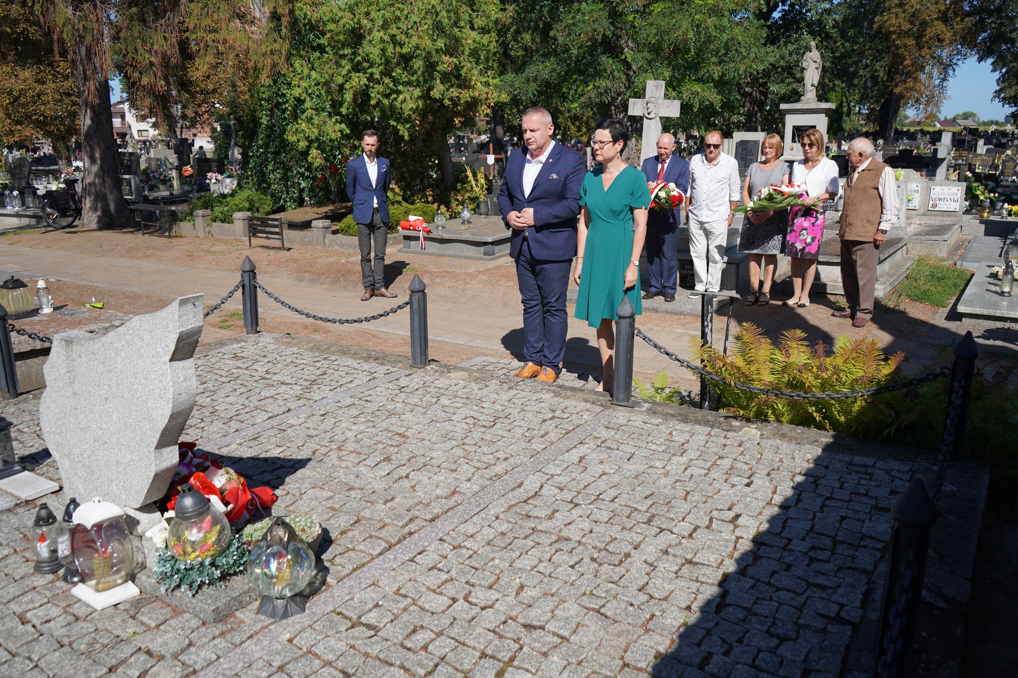 Na zdjęciu widzimy grupę ludzi składającą kwiaty i zapalające znicze przy grobie na cmentarzu. Uczestnicy stoją w skupieniu, oddając hołd zmarłemu. Na pierwszym planie nagrobek otoczony kwiatami i zniczami. Tło stanowi zadrzewiony teren