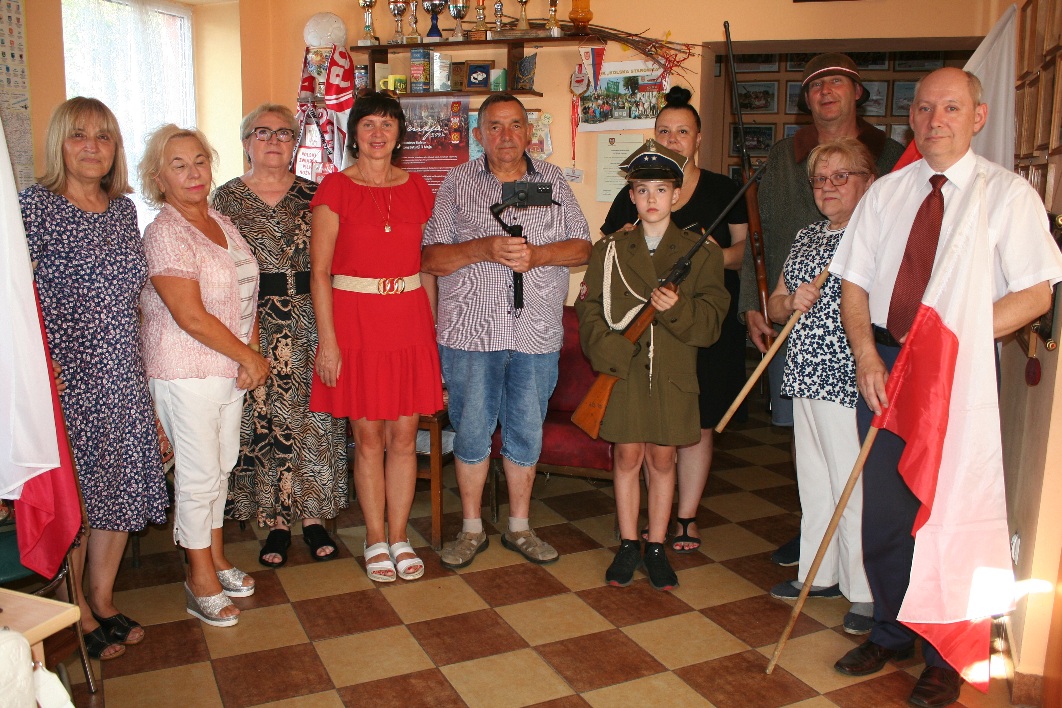 Na zdjęciu znajduje się grupa dziesięciu osób, w różnym wieku, stojących wewnątrz pomieszczenia. W tle widoczne są półki z pucharami i różnymi przedmiotami. Kilka osób trzyma flagi Polski, a jedna osoba (dziecko) ubrana jest w mundur wojskowy.