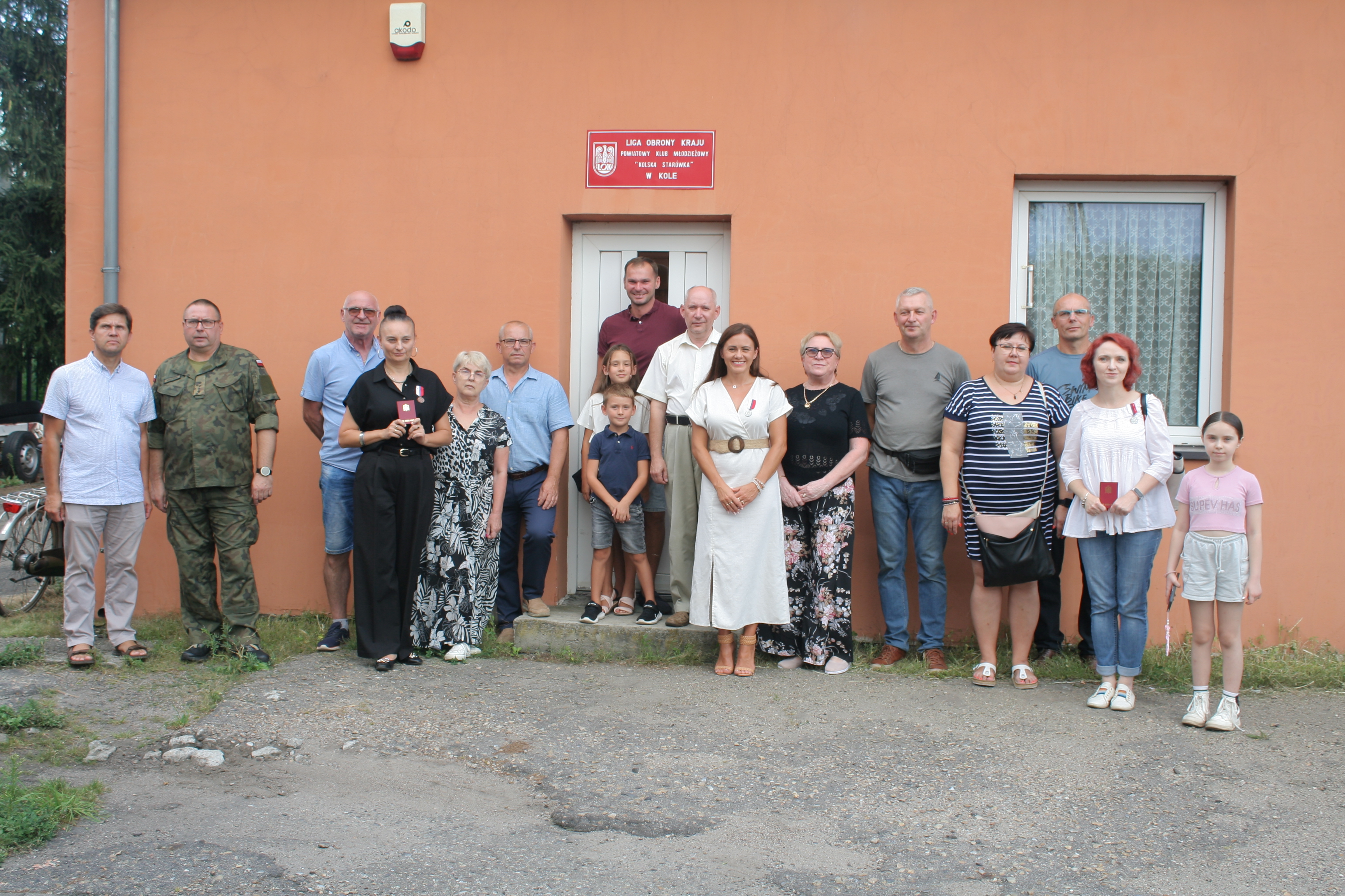 Na zdjęciu widzimy grupę ludzi stojących przed budynkiem, na którym znajduje się tablica z napisem LIGA OBRONY KRAJU. W środku grupy znajduje się kobieta w białej sukni, obok niej są dzieci oraz kilka innych osób. 