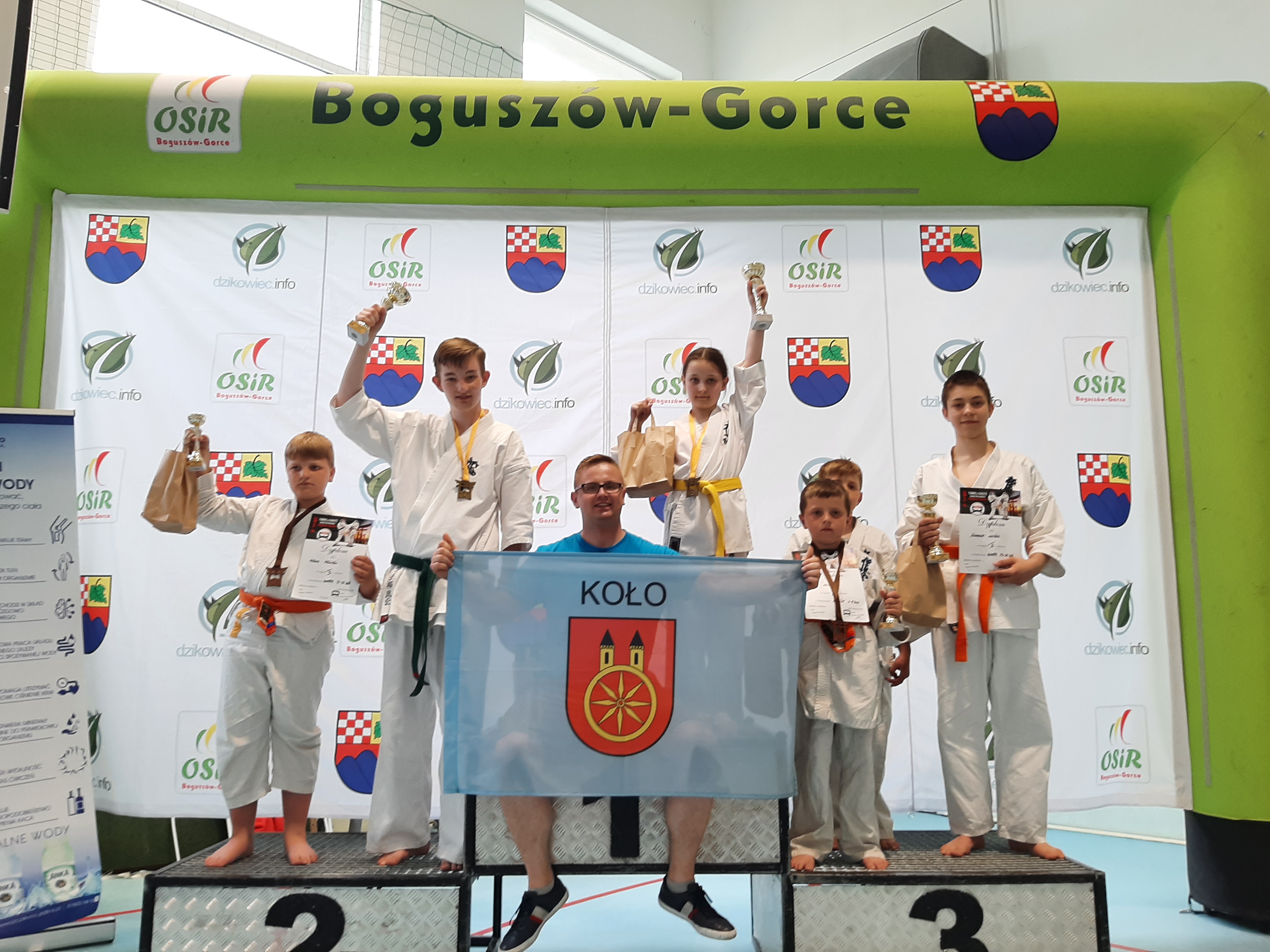 Kolscy karatecy Kolski Klub Kyokushin Karate stoją na podium, zawodnicy trzymają flagę miasta Koła.