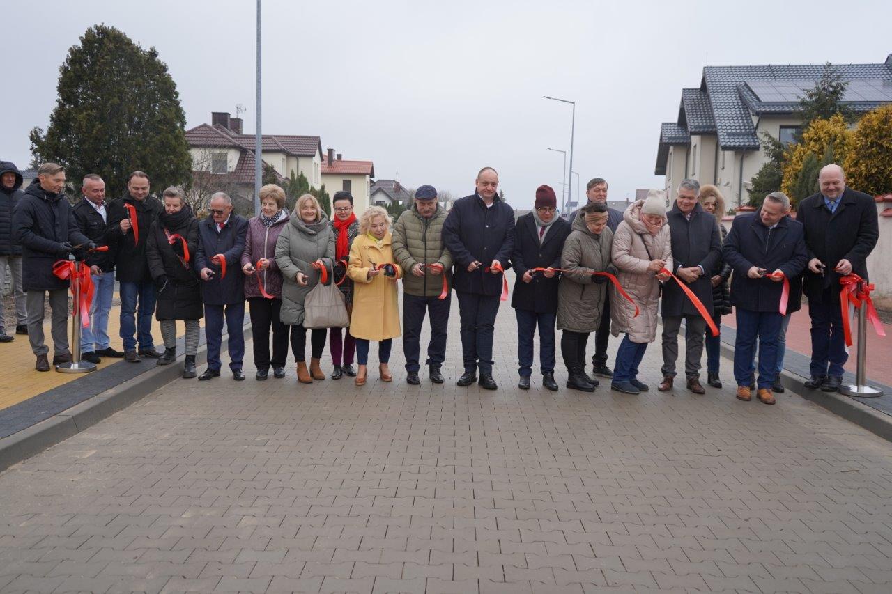 Na zdjęciu widać grupę osób biorących udział w uroczystym otwarciu drogi w Kole. Uczestnicy przecinają wstęgę. W tle są widoczne budynki mieszkalne oraz latarnie uliczne. Osoby są ciepło ubrani w kurtki i płaszcze.