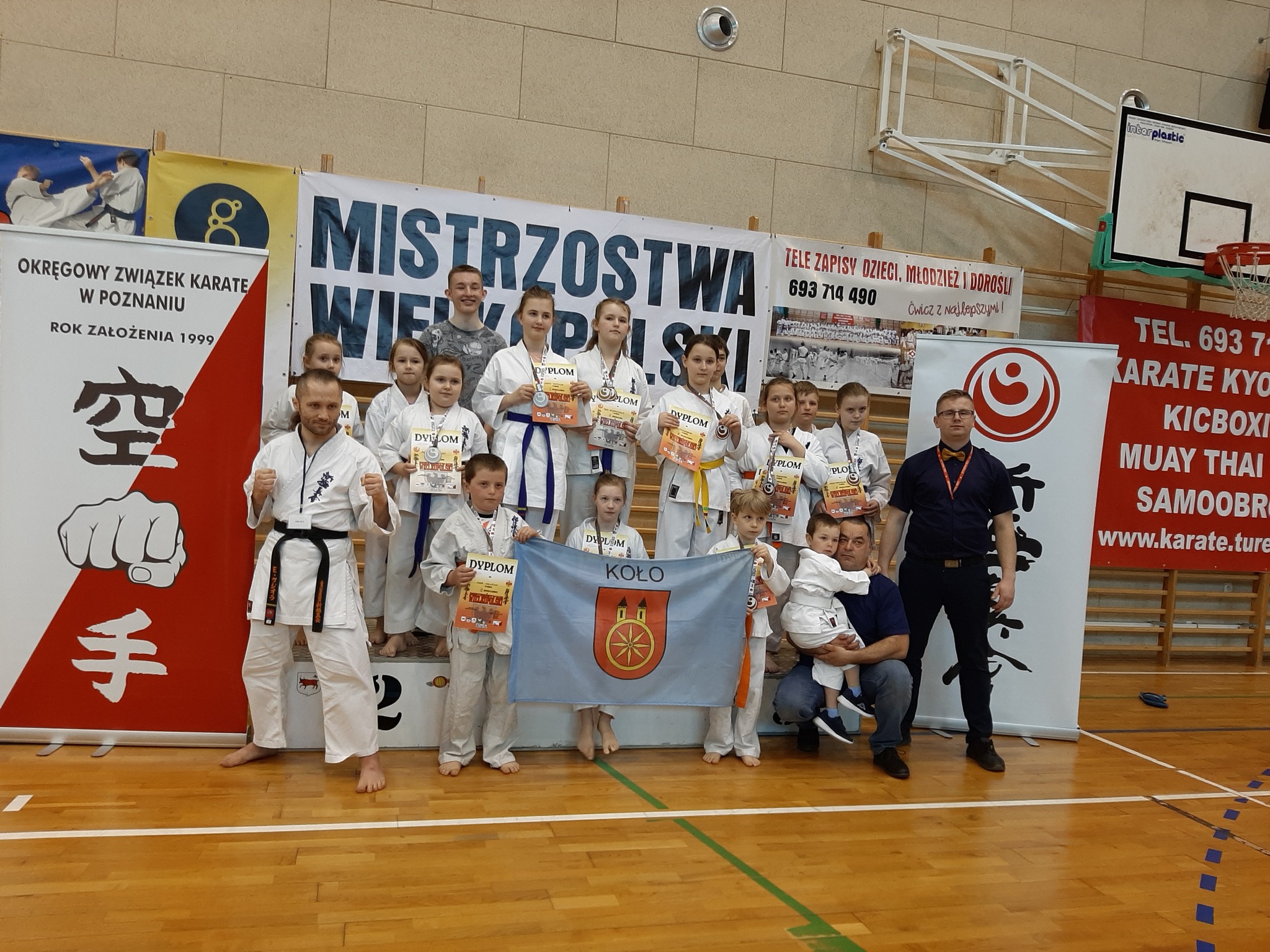 Kolscy karatecy Kolski Klub Kyokushin Karate stoją na podium, zawodnicy trzymają flagę miasta Koła.