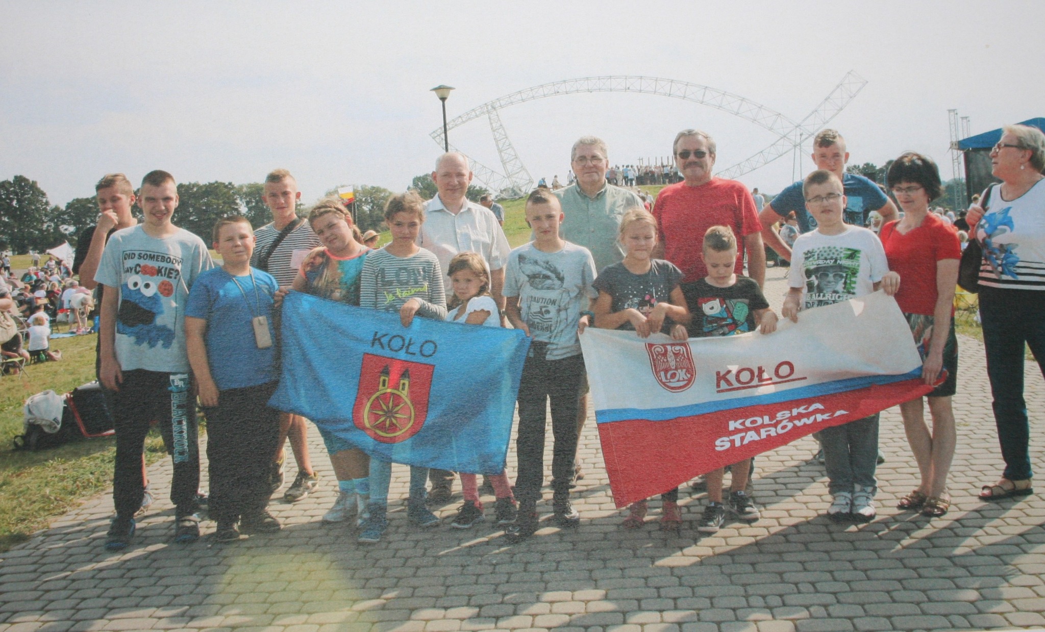 Na zdjęciu widać grupę osób, w tym dzieci, młodzież i dorosłych. Trzymają dwie flagi, jedna z flag jest niebieska z herbem Koła, druga biało-czerwona z napisem KOLSKA STARÓWKA. W tle widać duży, otwarty teren z metalową siecią symbolizującą bramę. 