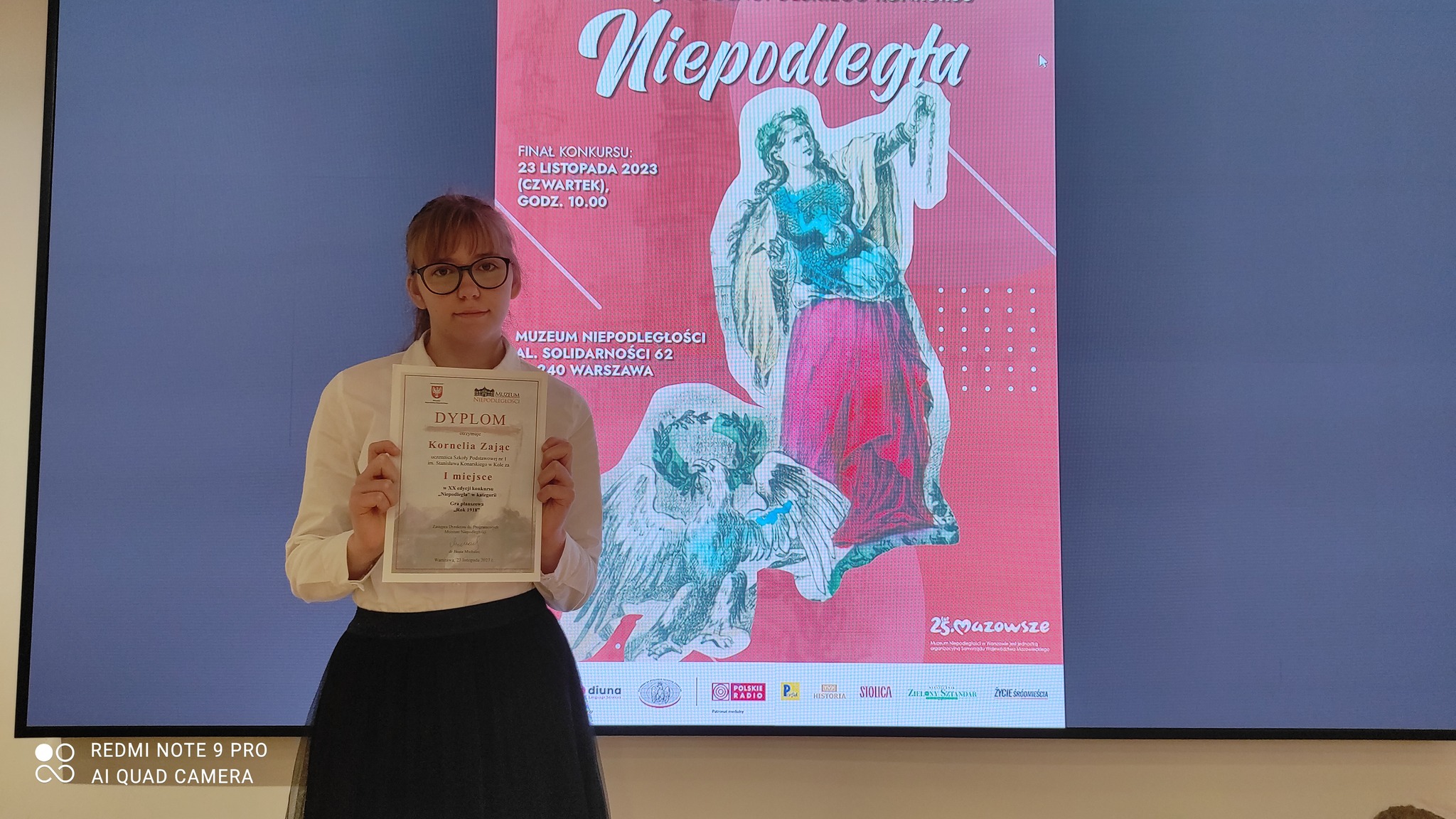 Kornelia Zając z dyplomem w dłoniach, w tle plakat XX edycji Ogólnopolskiego Konkursu „Niepodległa” 
