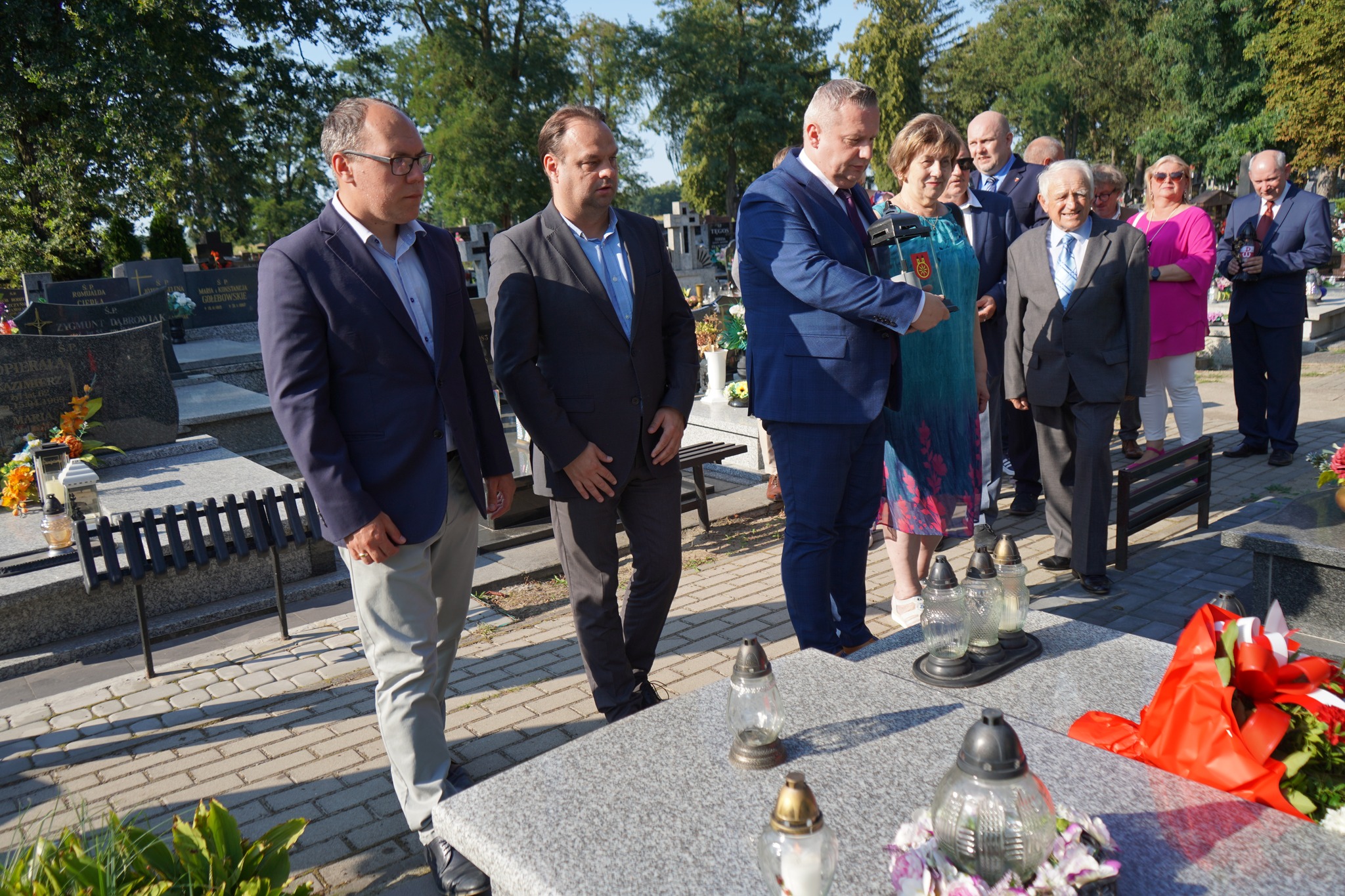 Zdjęcie przedstawia grupę ludzi zgromadzonych na cmentarzu. W tle widać zielone drzewa, a grupa osób trzyma znicze. Grób jest udekorowany kwiatami i zniczami.