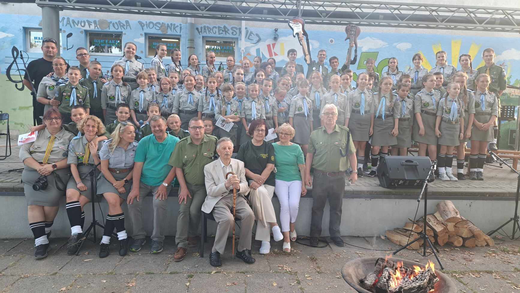 Zdjęcie przedstawia dużą grupę harcerzy i dorosłych, którzy pozują wspólnie na scenie na tle malowidła ściennego z napisem „Manufaktura Piosenki Harcerskiej”. Na pierwszym planie widać ognisko.