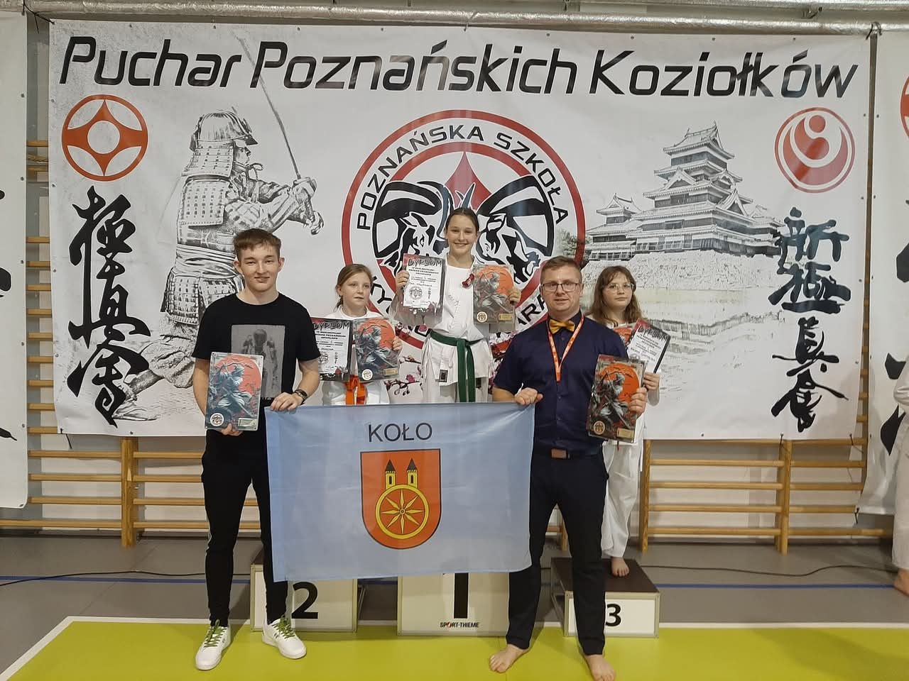 Na zdjęciu znajduje się grupa osób stojących na podium podczas Pucharu Poznańskich Koziołków. Centralnie widać zwyciężczynię na najwyższym stopniu podium w stroju karate, trzymającą dyplom i nagrody. Obok niej inne zawodniczki oraz mężczyzna z flagą 