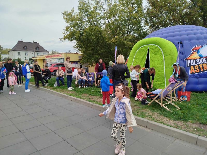 Na zdjęciu widać grupę osób, głównie rodzin z dziećmi, w plenerze. W tle znajduje się dmuchana kopuła planetarium oznaczona napisem Polskie Astro oraz charakterystycznym logiem z astronautą, niektórzy siedzą na leżakach Dzieci bawią się na trawie