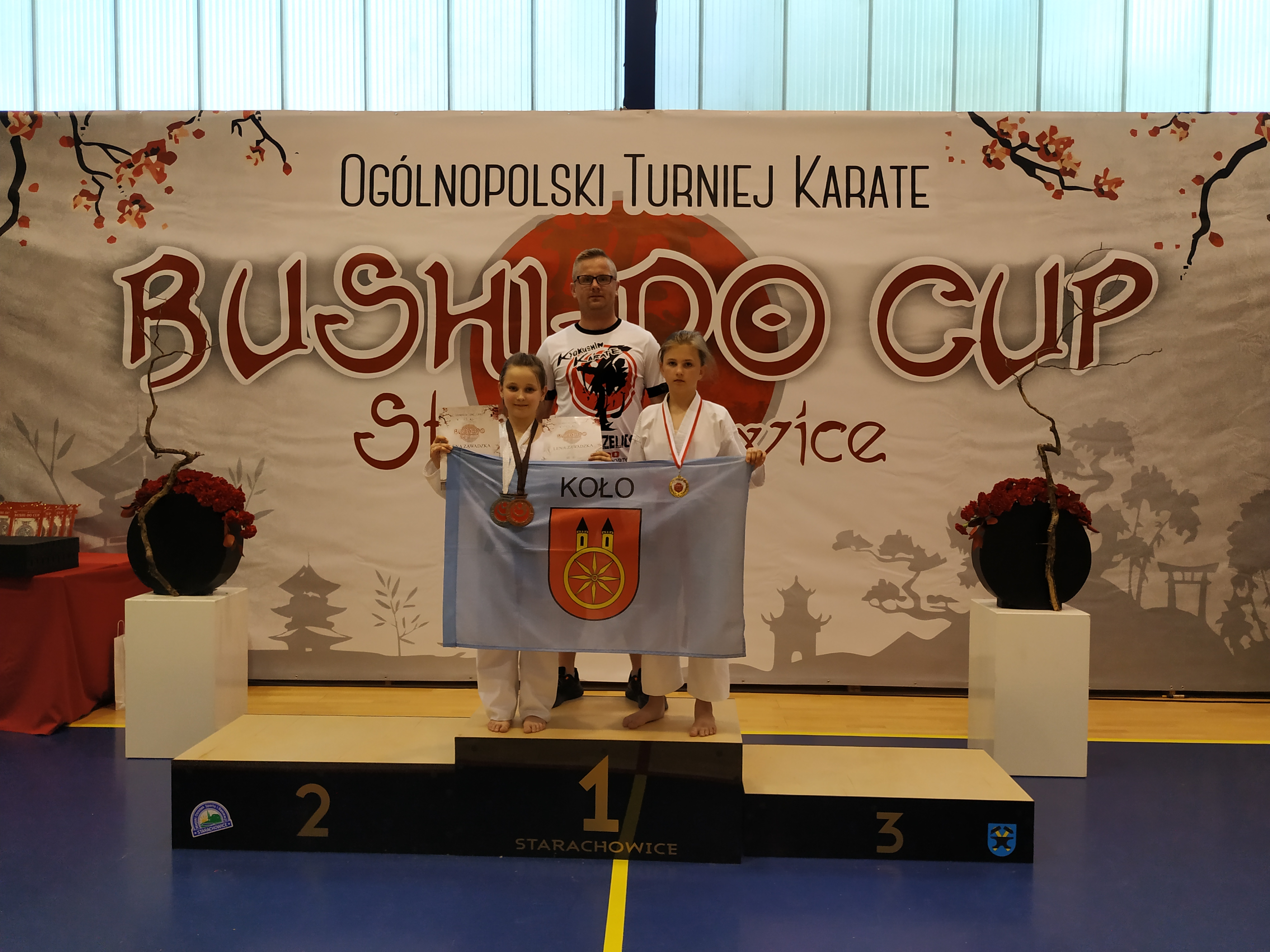 Lena Zawadzka i Nadia Antecka stoją na podium z medalami i flagą miasta Koła z opiekunem.