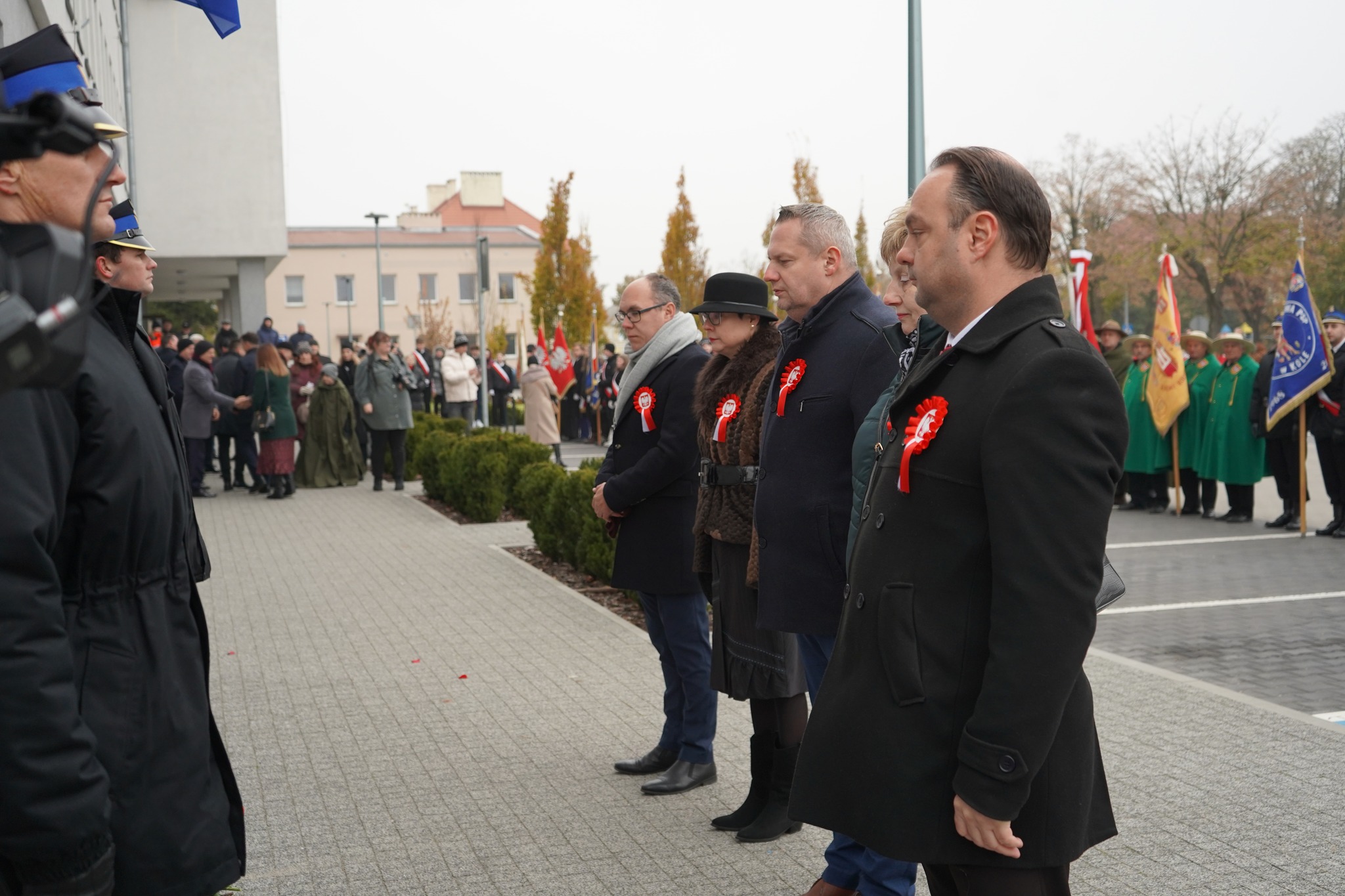 Na zdjęciu widzimy delegację władz miasta Koła w oficjalnych obchodach Narodowego Święta Niepodległości. Ubrani są elegancko z biało-czerwonymi kotylionami, stoją w szeregu, zachowując powagę i szacunek. W tle grupa osób oraz poczty sztandarowe.