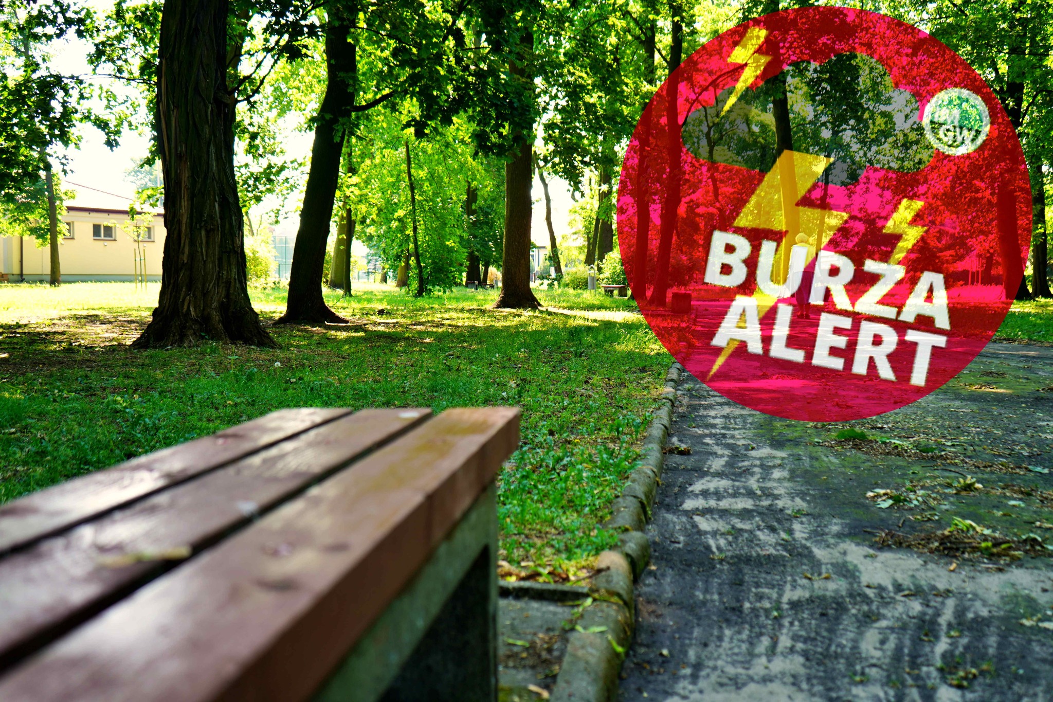Zdjęcie przedstawia park w słoneczny dzień, z trawiastą przestrzenią, drzewami i ścieżką spacerową. Na pierwszym planie znajduje się ławka. Na zdjęciu nałożony jest czerwony okrągły znak z napisem BURZA ALERT.