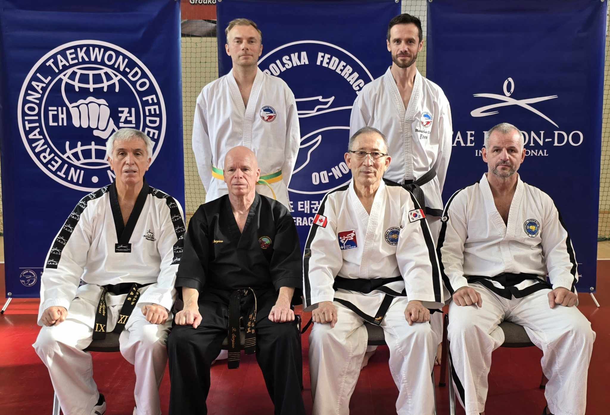 Na zdjęciu znajduje się sześciu mężczyzn ubranych w stroje do taekwondo, z umieszczonymi emblematami i stopniami na pasach. Trzech z nich siedzi z przodu, a troje stoją z tyłu. Na drugim planie banery z logo różnych organizacji związanych z taekwondo