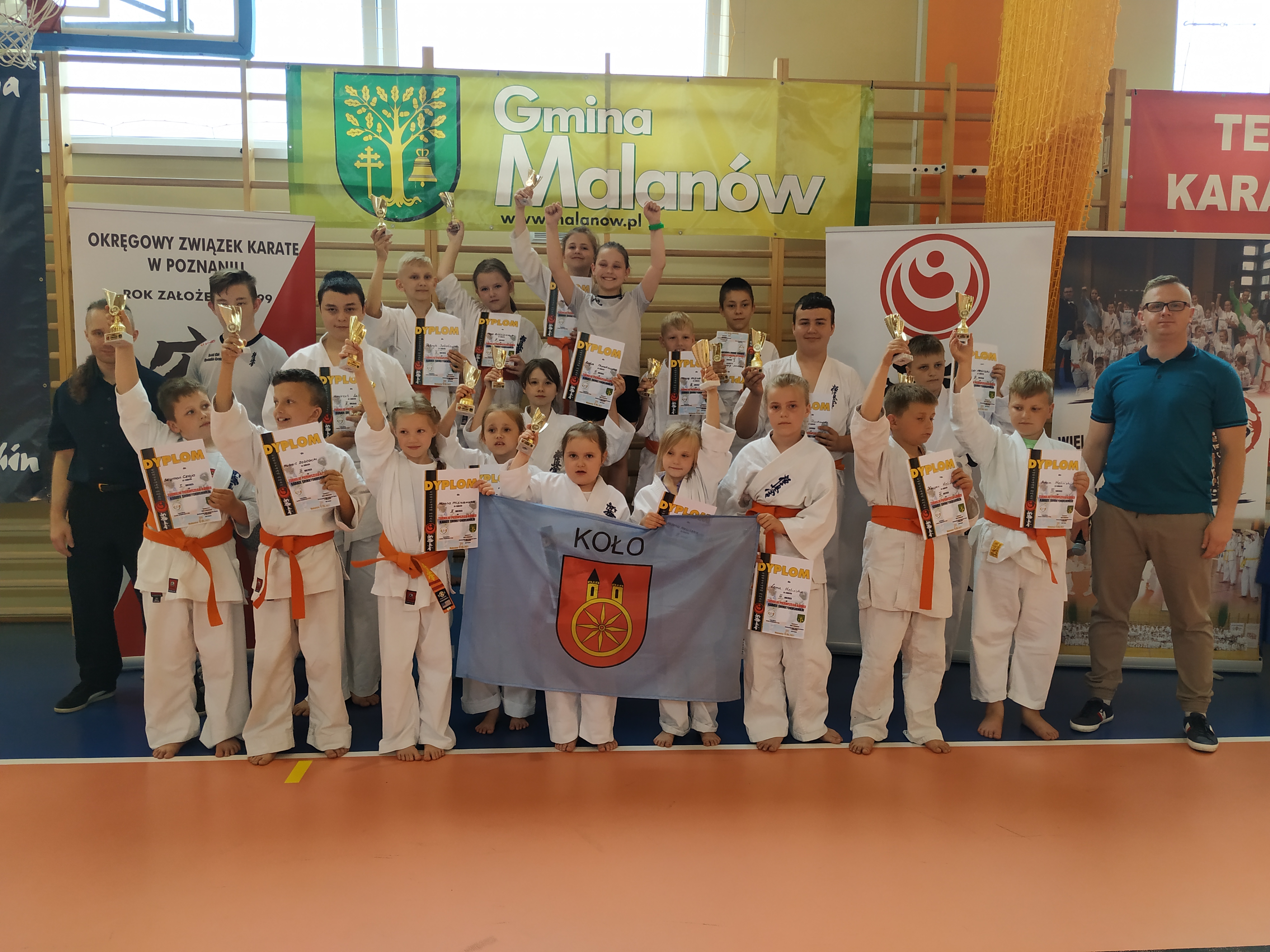 Grupowe zdjęcie kolskich zawodniczek i zawodników z dyplomami i pucharami wraz z opiekunami.